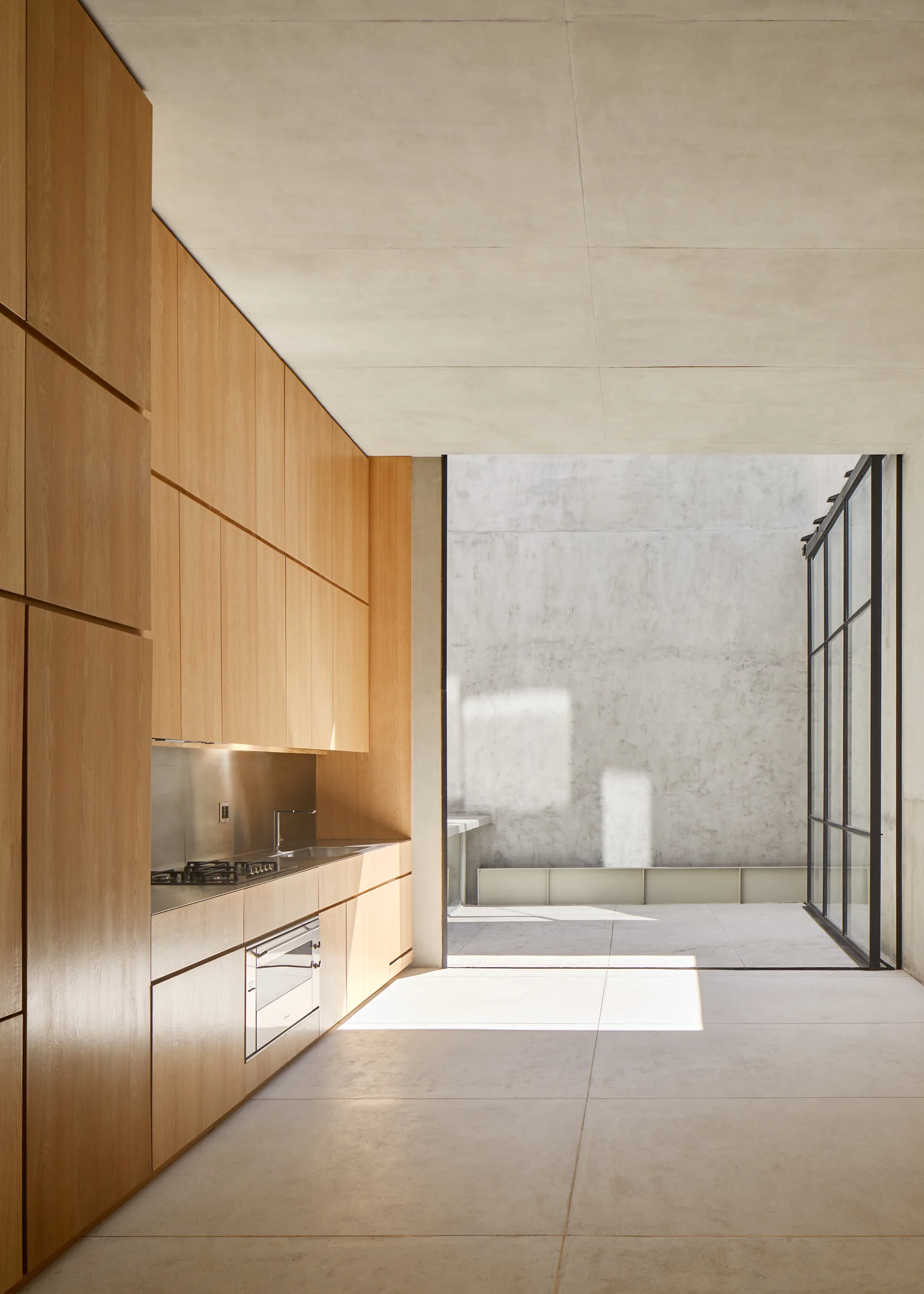 A minimalist kitchen featuring warm wood cabinetry and a stainless steel countertop opens to an outdoor concrete courtyard through a tall glass door.