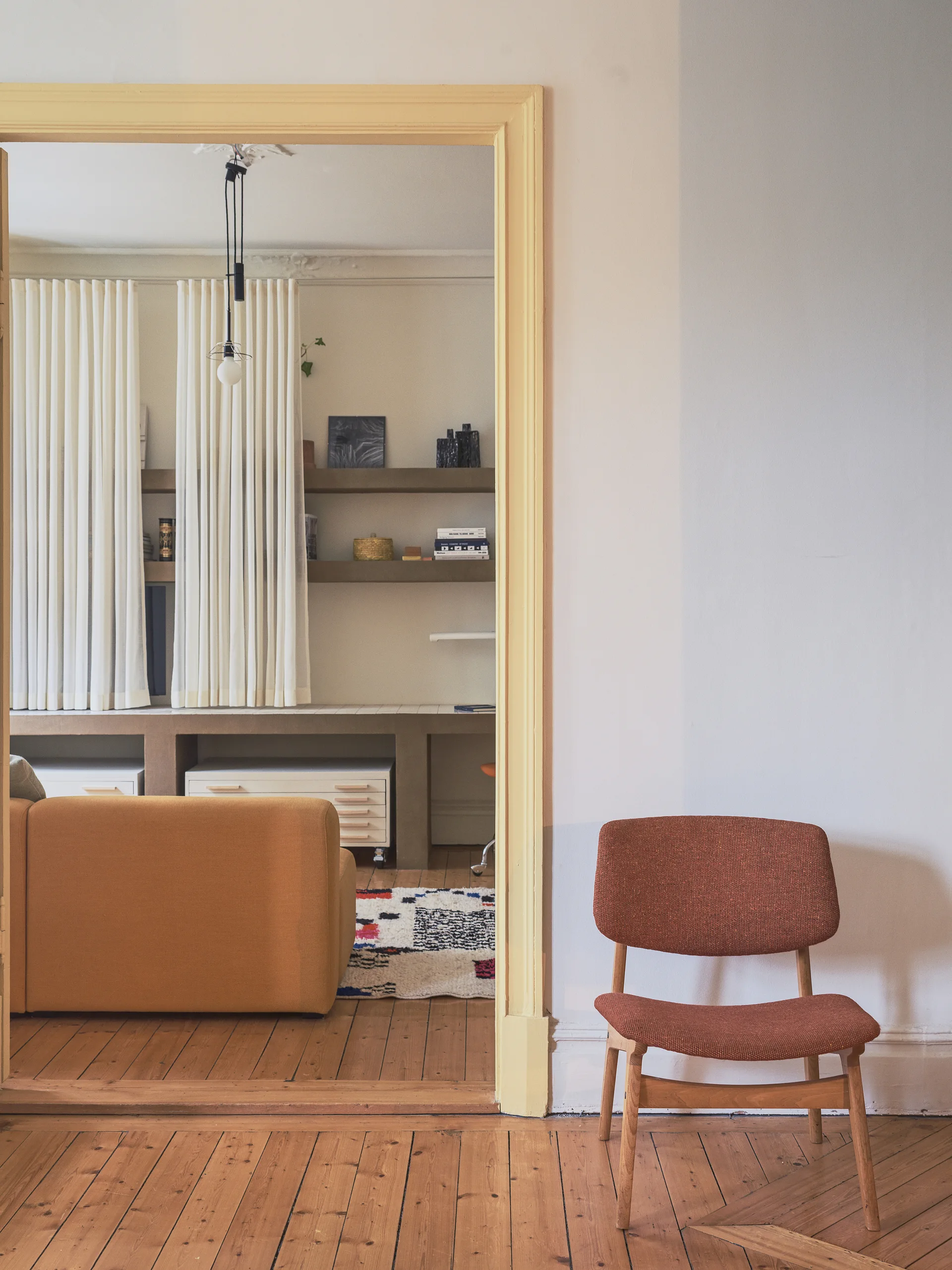A rust colored wooden chair sits on wood flooring next to a yellow painted doorway leading into a living room with an orange sofa.