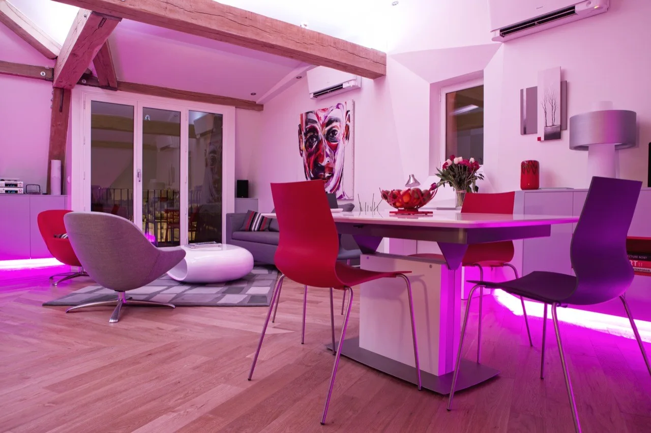A modern open-plan dining and living area featuring a white table with red chairs, rustic wood ceiling beams, and a vibrant pink floor-level LED glow.