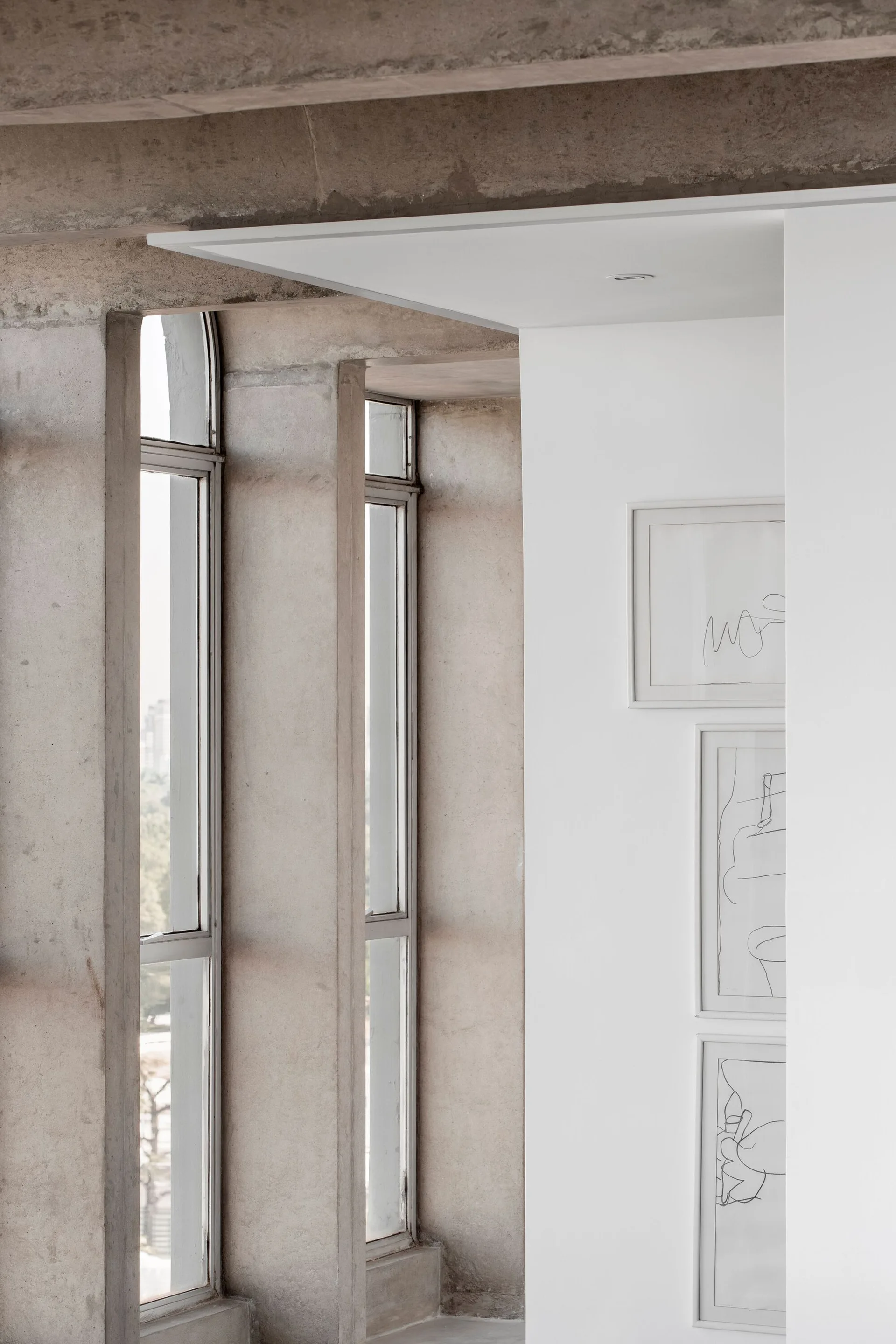 Tall narrow windows set in rough concrete walls next to a smooth white partition wall hanging three framed minimalist line drawings.