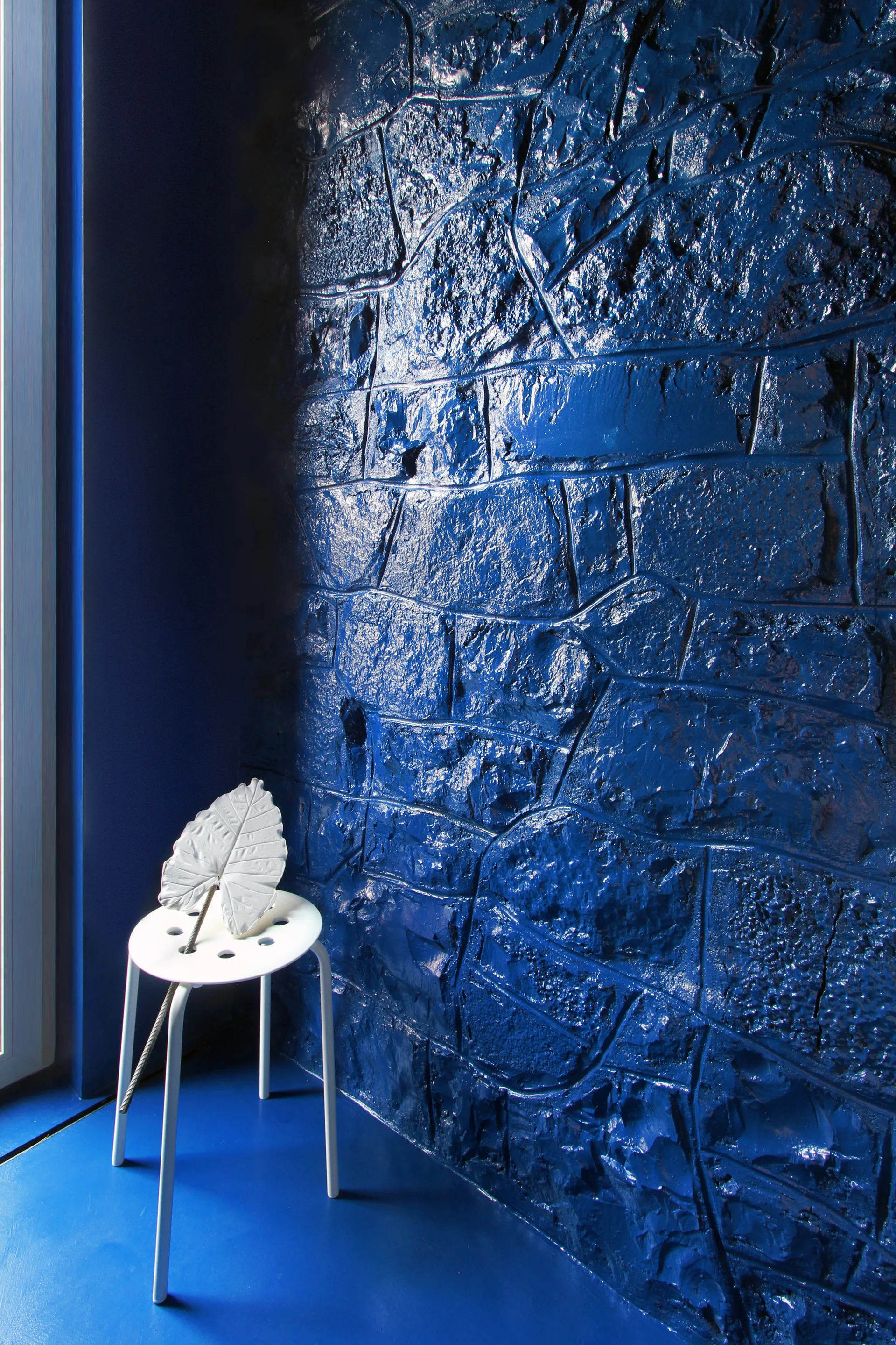 A minimalist white metal stool stands on a blue floor in front of a high-gloss, craggy blue stone accent wall with a leaf sculpture.