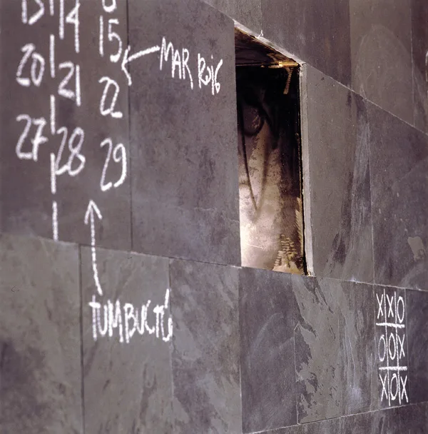 A close-up view of dark grey slate wall tiles featuring a small square recessed niche and white chalk drawings and numbers.