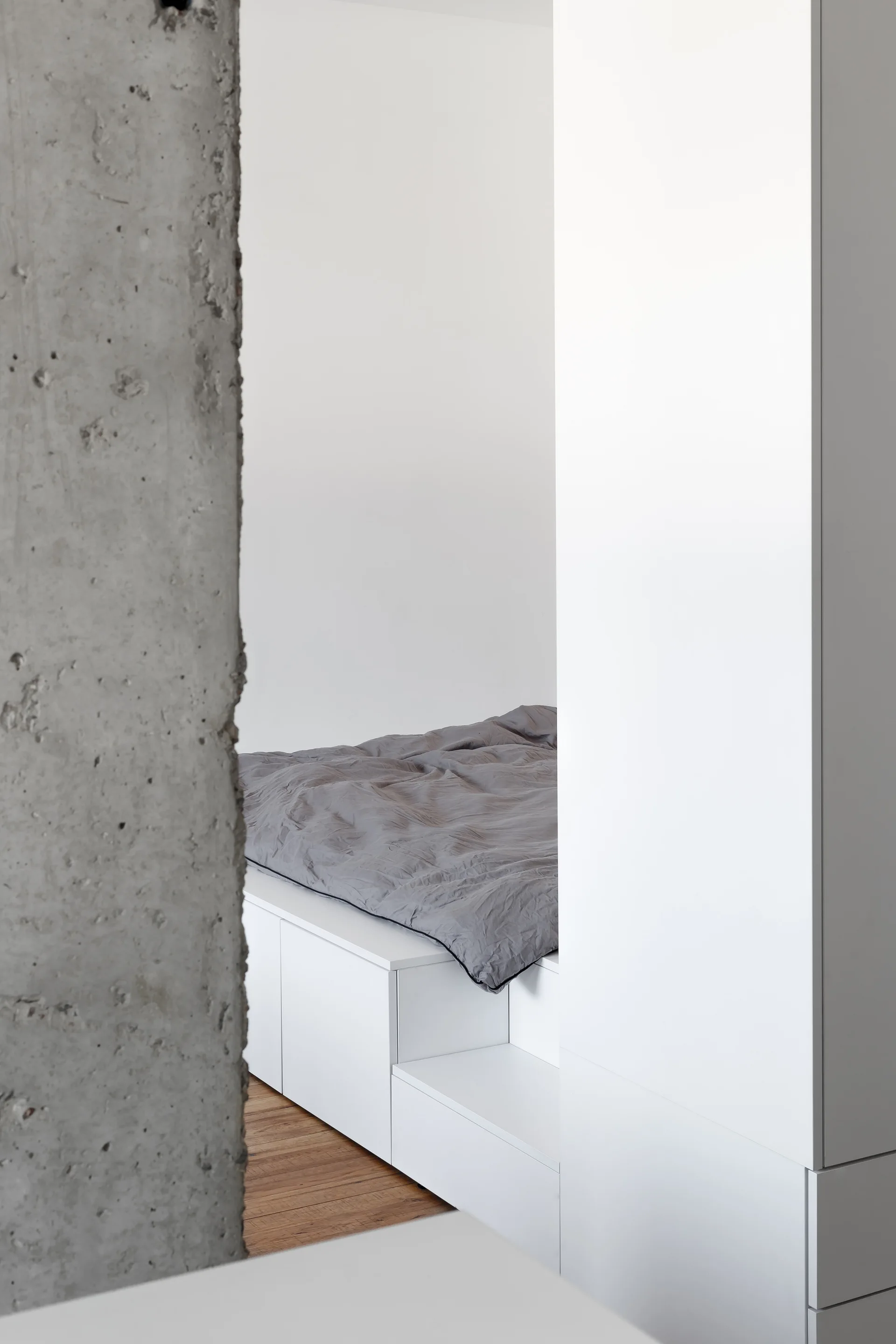 A minimalist platform bed with grey bedding is recessed between a clean white built-in storage unit and a textured raw concrete pillar.