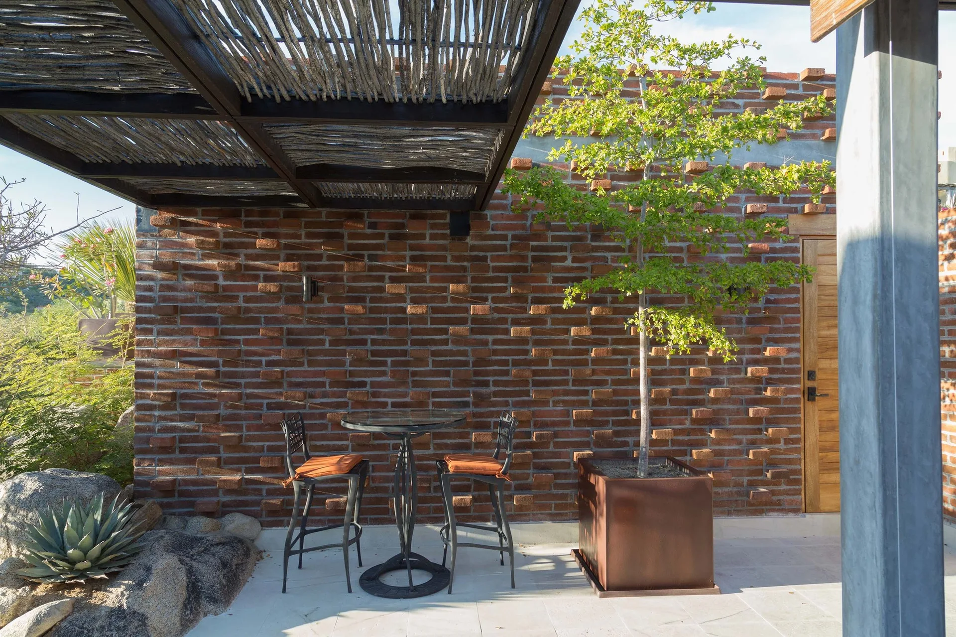 An outdoor patio features a black metal bistro set and a potted tree against a textured red brick wall under a stick-roof pergola.