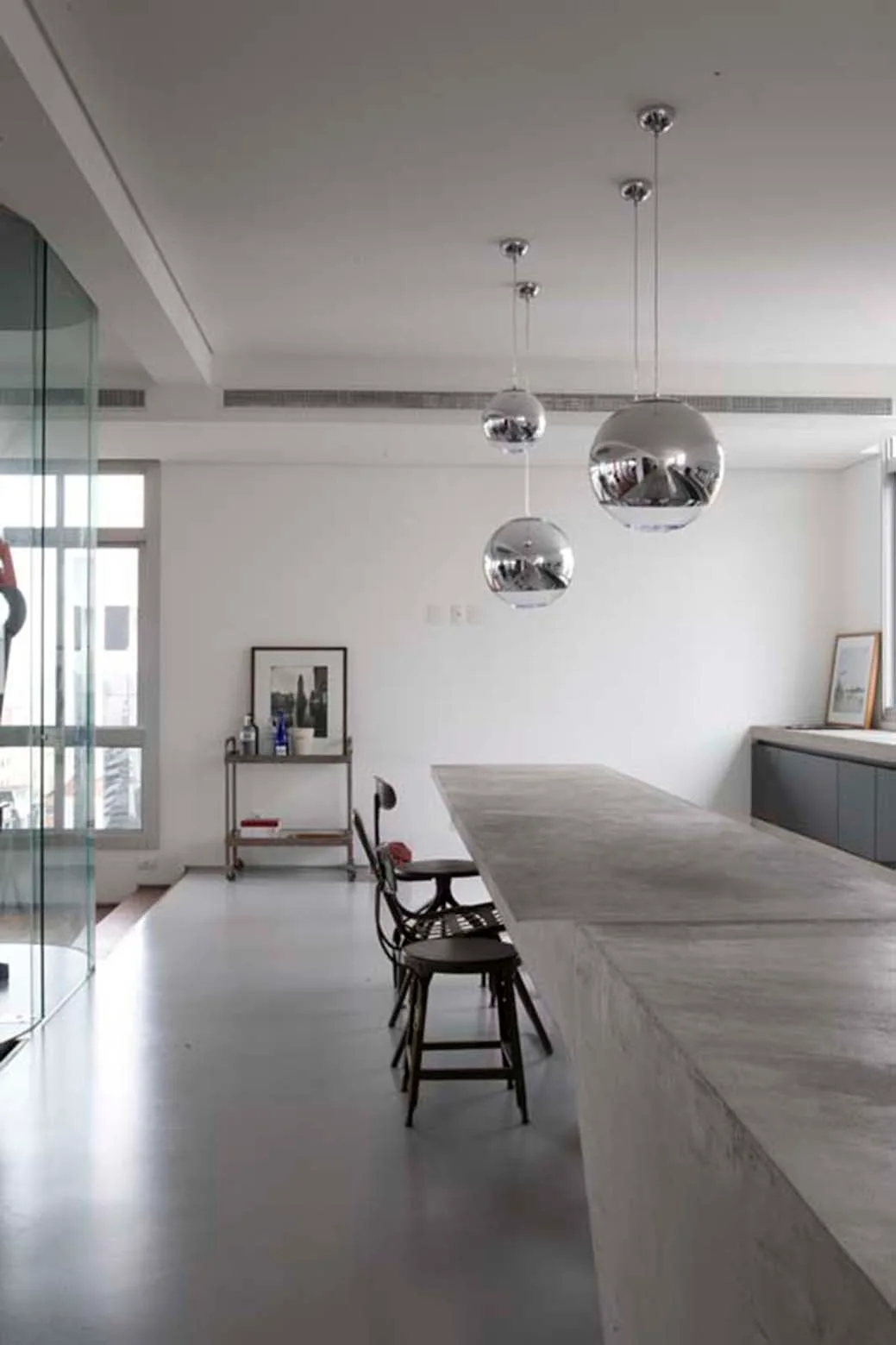 A minimalist kitchen features a large concrete island with two dark bar stools beneath three reflective chrome spherical pendant lights.