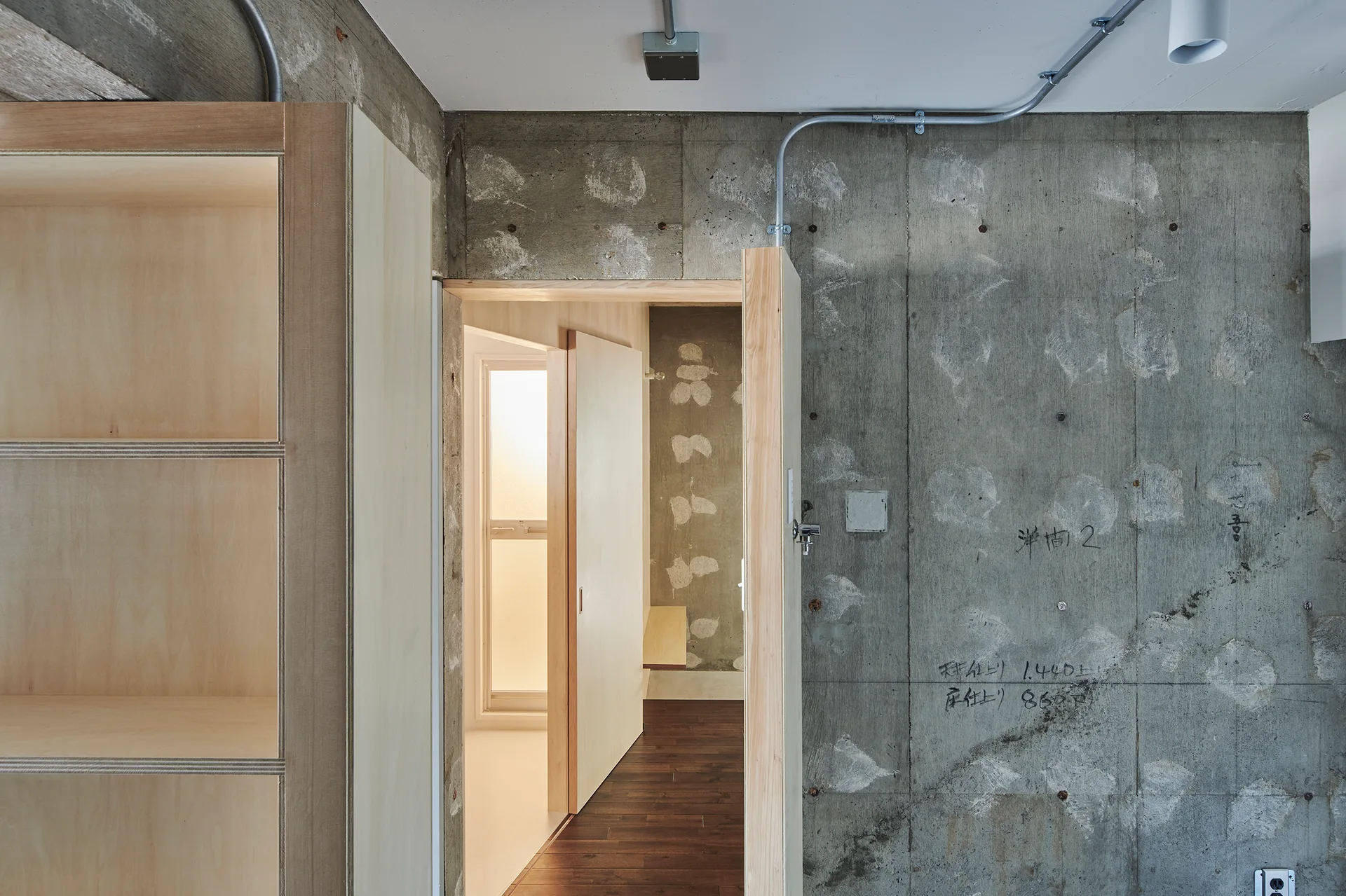 An industrial entryway features raw concrete walls with construction markings, exposed electrical conduit, and a light-toned plywood cabinet.
