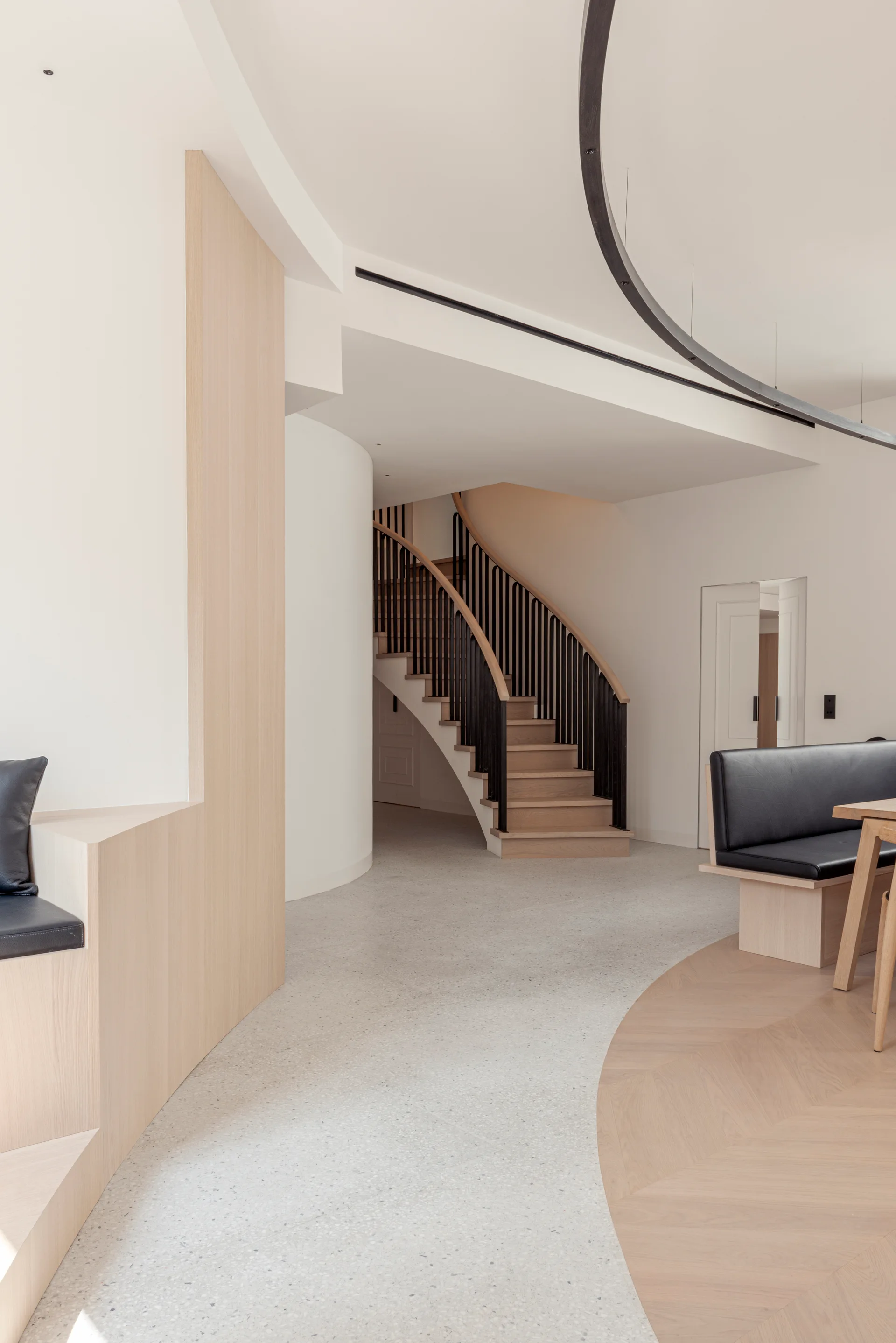 A minimalist hallway features a curved wooden staircase with black metal railings, terrazzo flooring, and integrated light wood banquette seating.