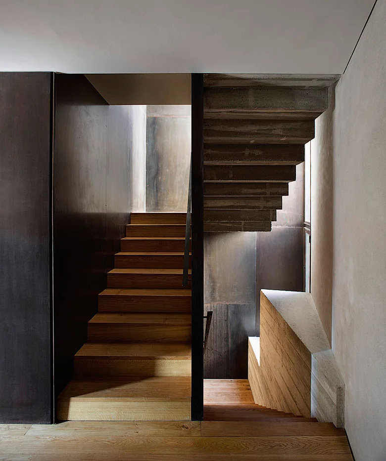 A minimalist stairwell featuring natural wood treads and an exposed raw concrete staircase next to dark wall paneling.