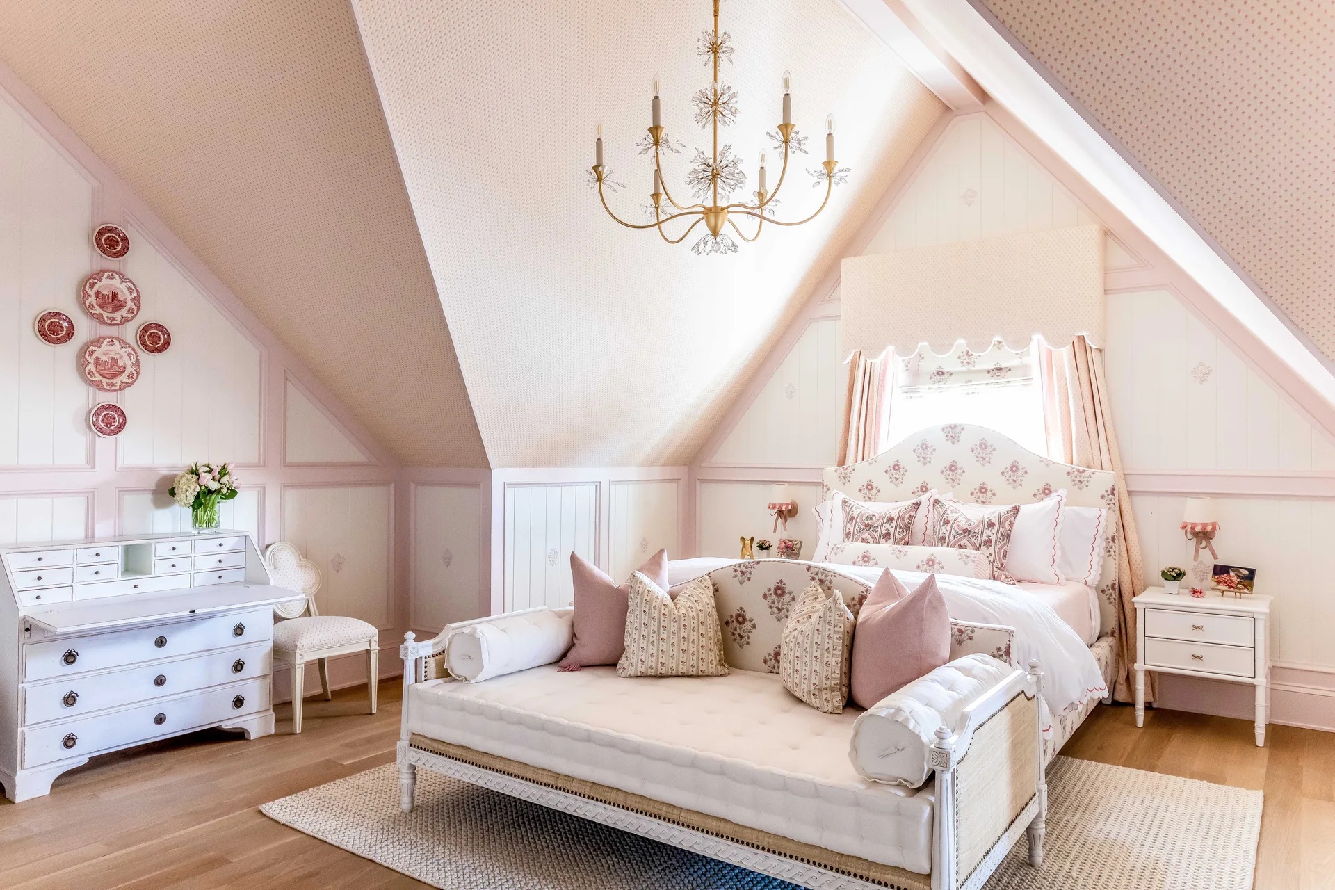 A bright attic bedroom with a daybed at the foot of an upholstered bed under a sloped pink patterned ceiling with a gold chandelier.