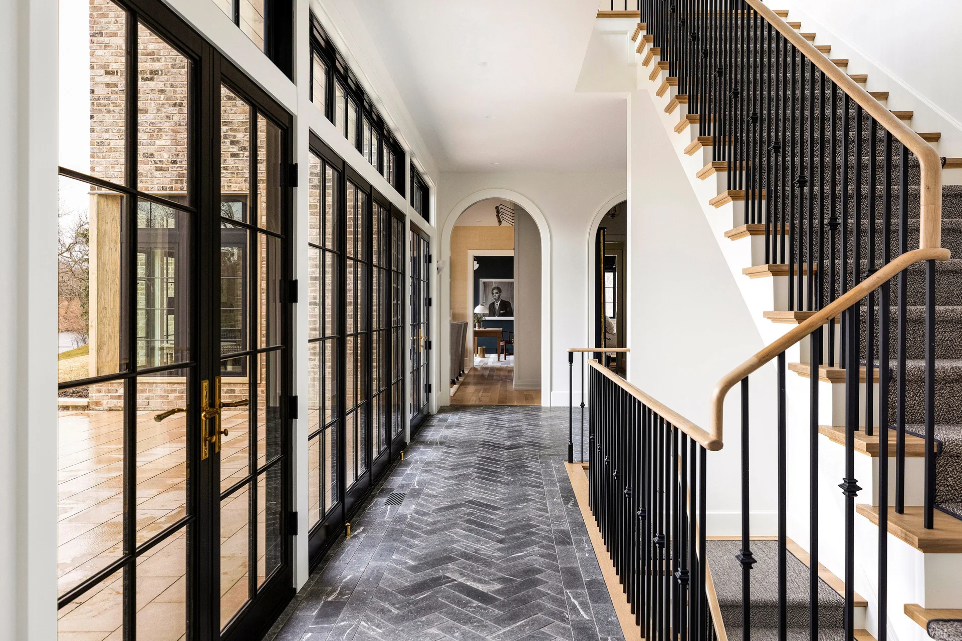 An interior hallway features a dark grey herringbone stone floor, black-framed glass patio doors, and a staircase with a black metal railing.