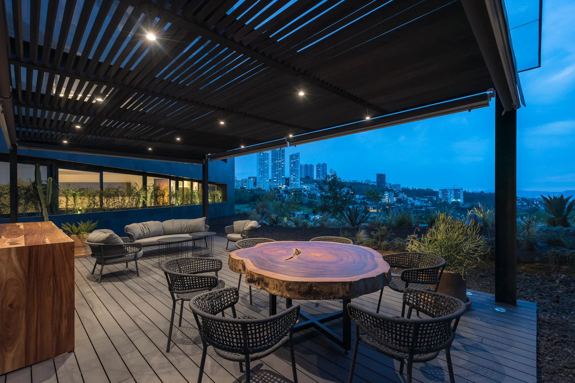 A twilight outdoor deck features a large live-edge wood dining table with woven chairs under a dark slatted pergola roof with recessed lighting.