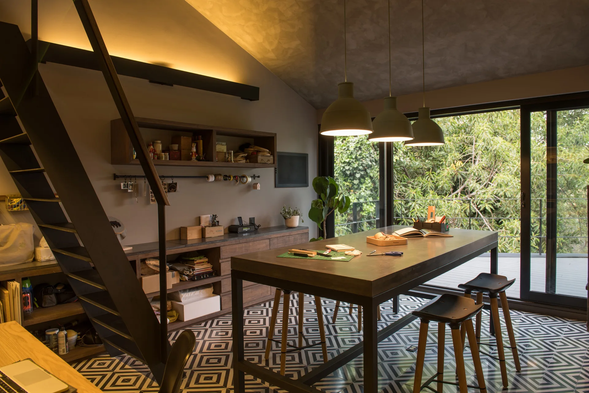 A modern home office features a long work table with stools, a metal staircase, wooden storage, and bold geometric floor tiles.