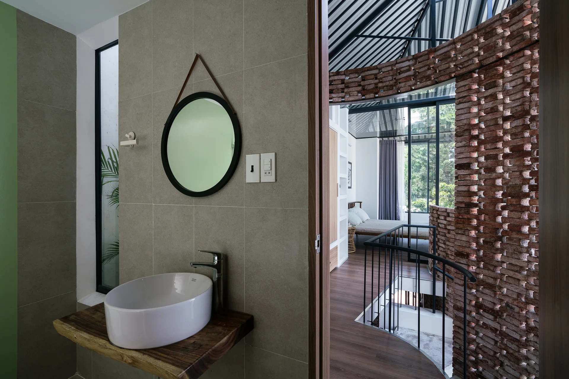 A modern bathroom with a white vessel sink on a live-edge wood vanity features a view into a bedroom with a curved red brick screen wall.