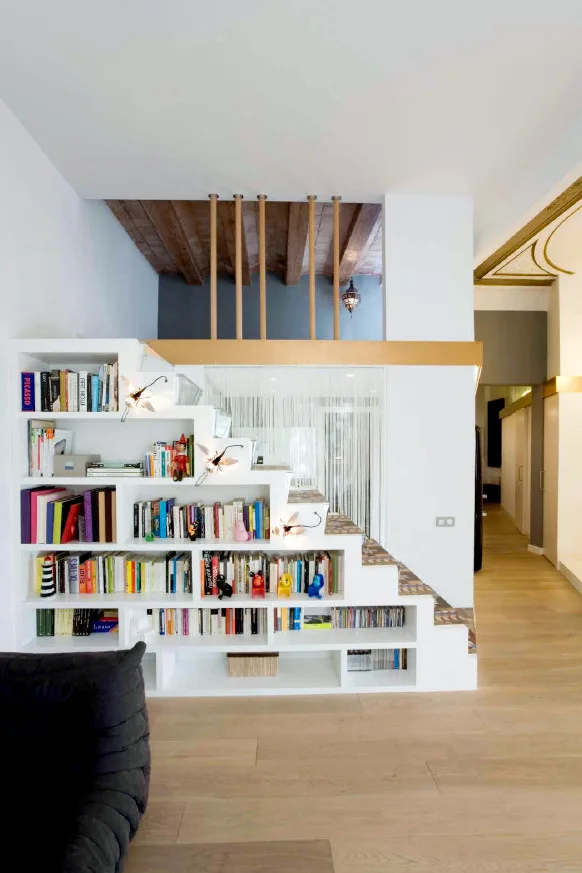 A custom white staircase built with integrated bookshelf storage stands on light wood flooring beneath exposed wooden ceiling beams.