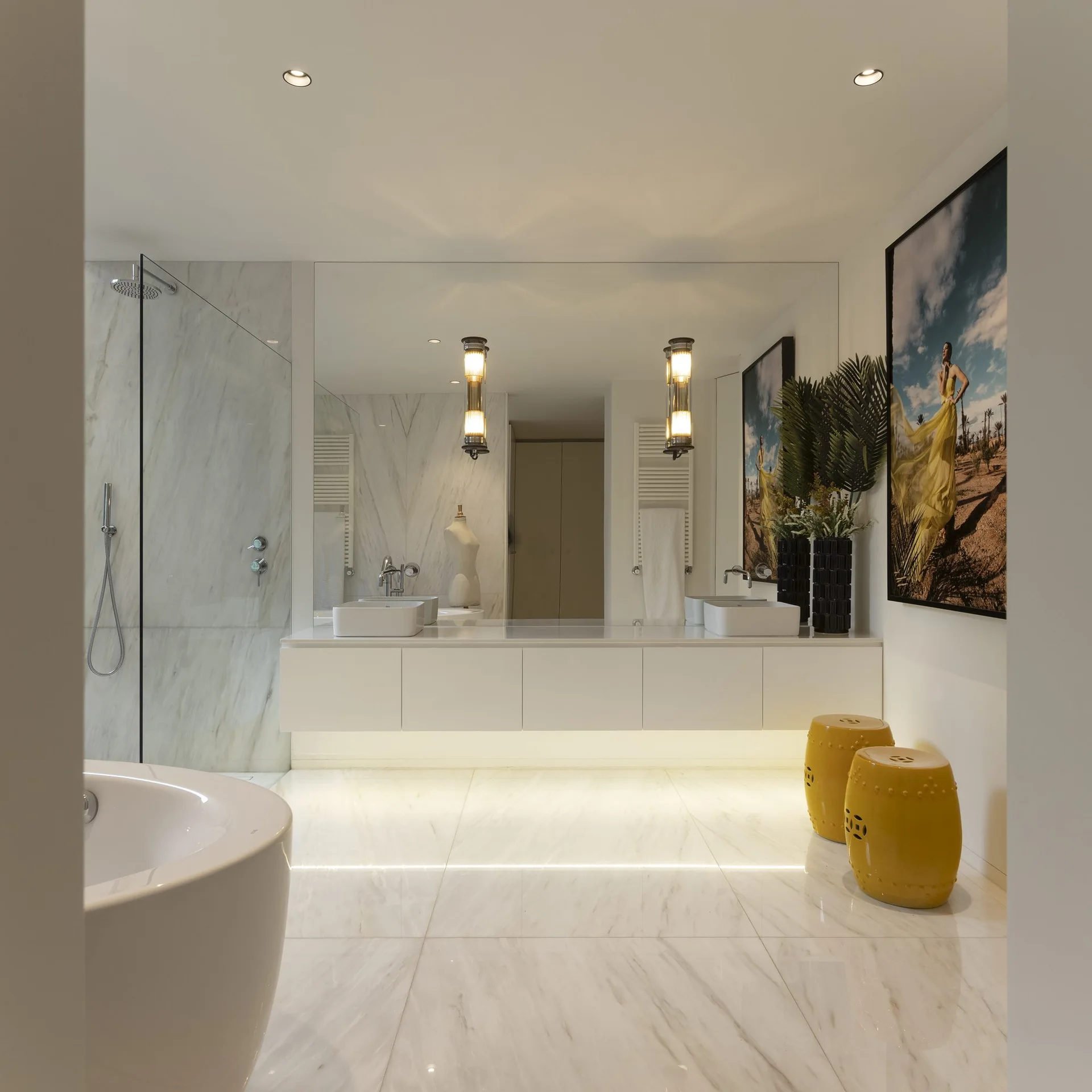 A bright bathroom features a floating white double vanity, large mirror, marble tile floor, and two yellow ceramic garden stools.