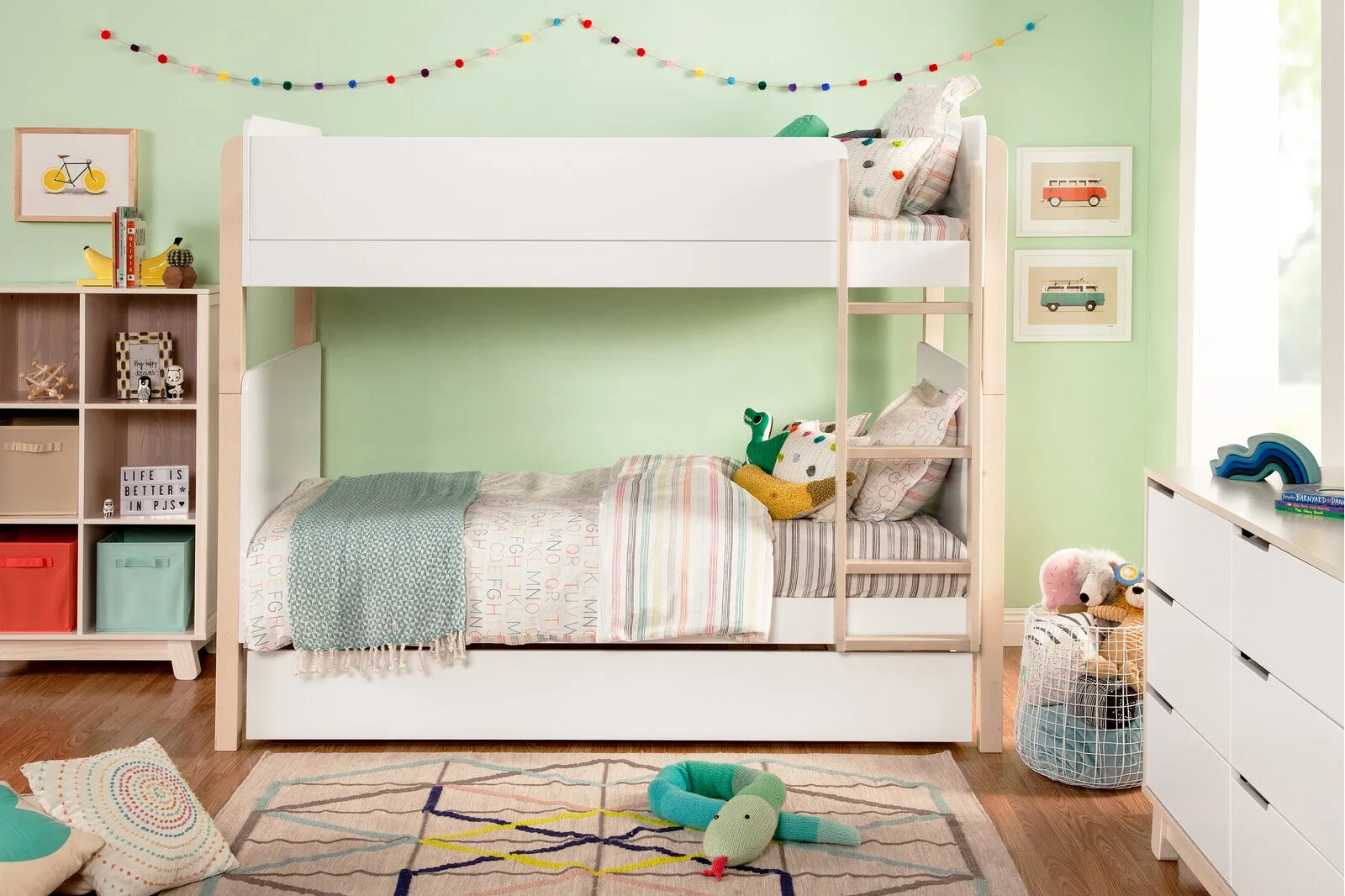 A white and wood bunk bed stands against a pastel mint green wall in a children's bedroom next to a bookshelf, dresser, and patterned rug.