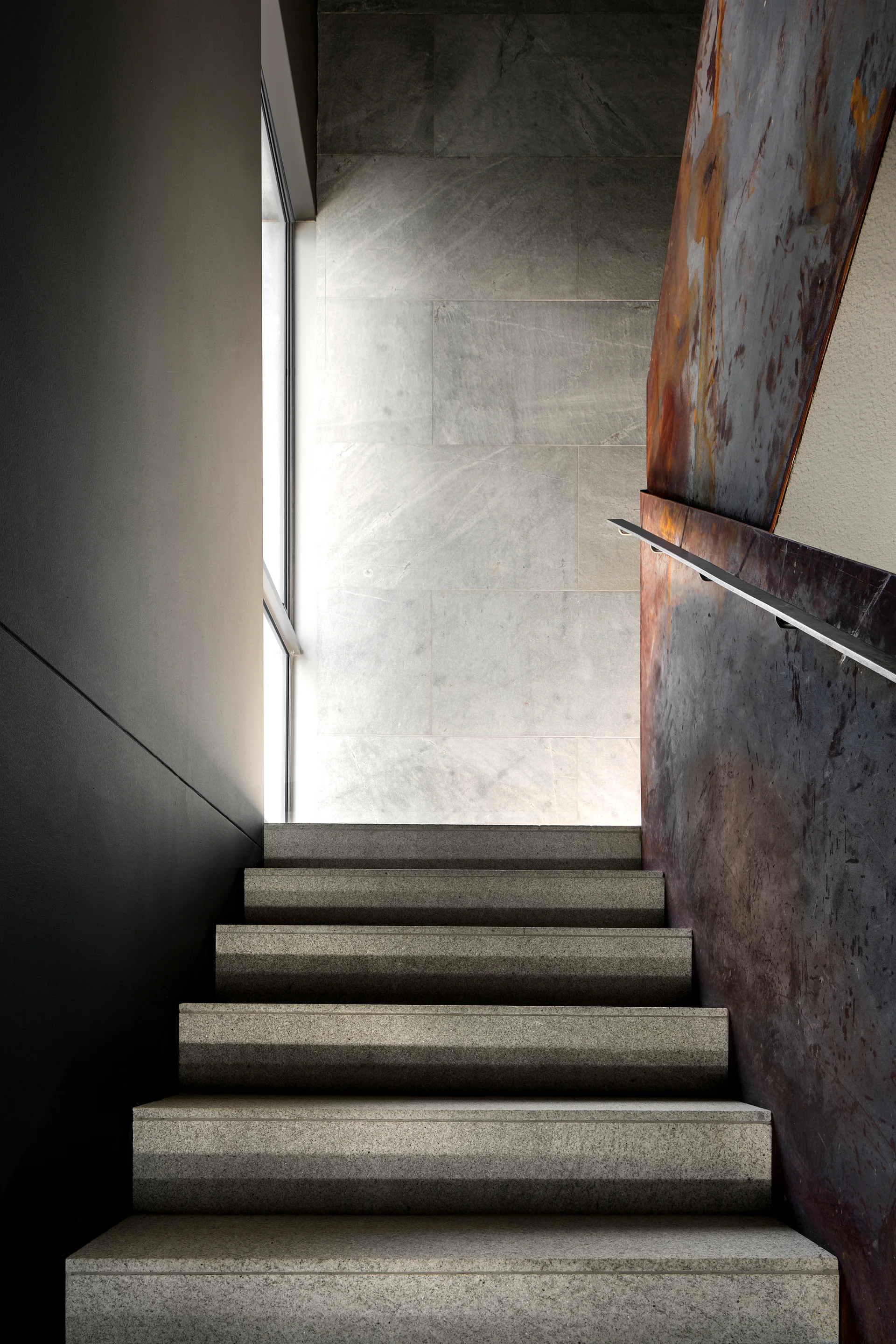 A modern stone staircase ascends between a dark grey wall and a weathered, rusted metal panel under natural light from a vertical window.