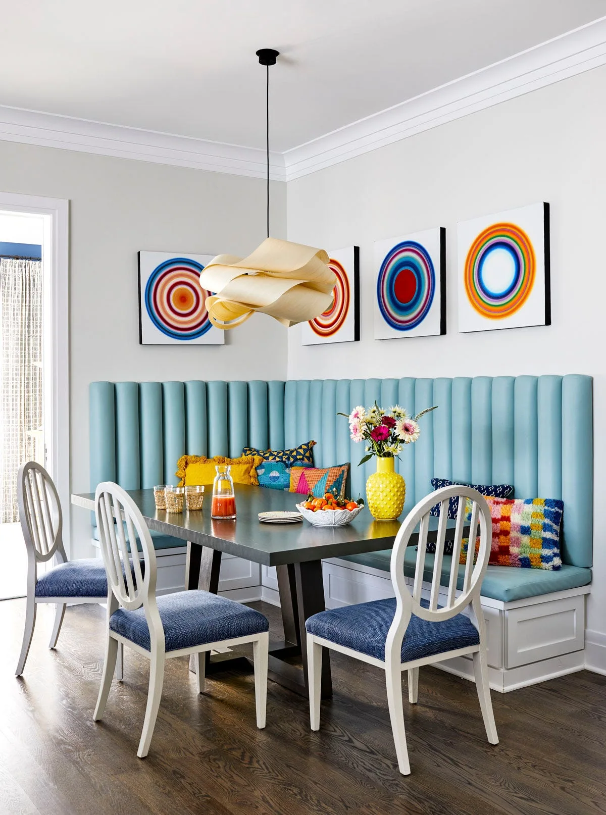 A dining nook features a light blue channel-tufted banquette bench, a dark wood dining table, white chairs with blue seats, and colorful wall art.