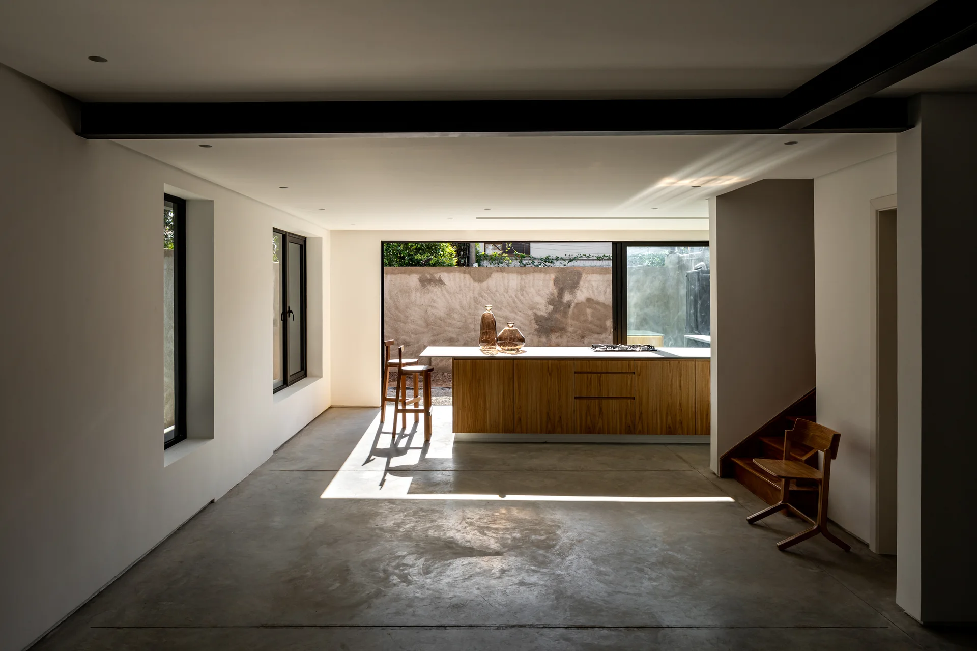 A minimalist kitchen features a wood-paneled island on a grey concrete floor beneath white walls and a dark structural steel beam.