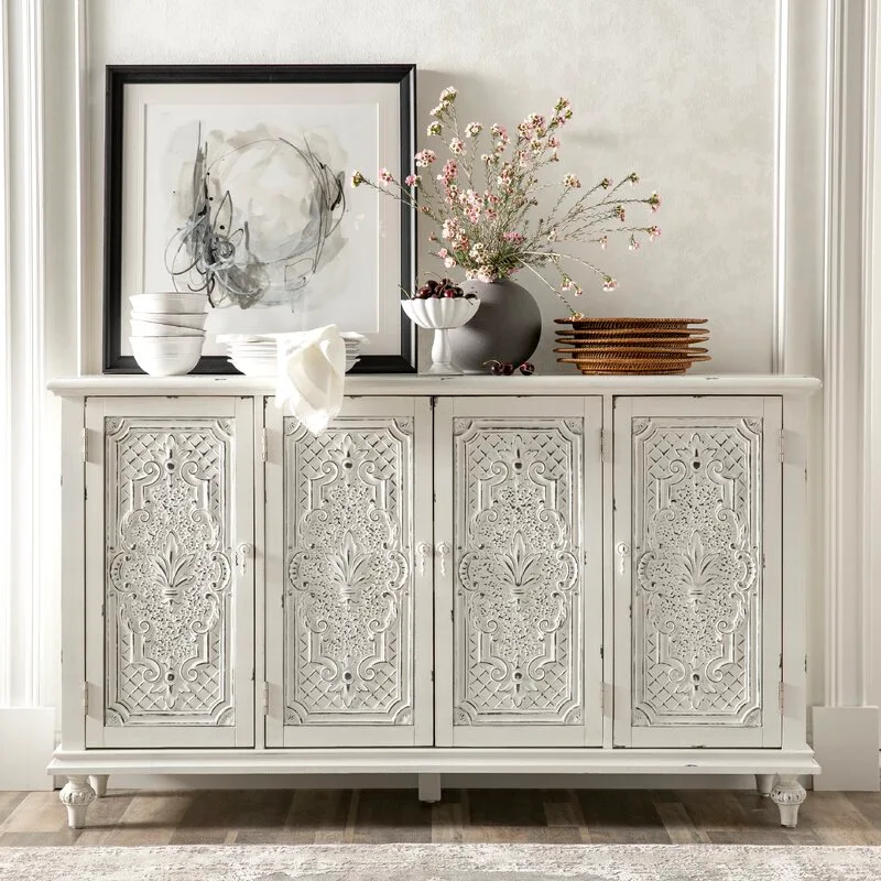 An ornate distressed white sideboard with relief-carved doors is decorated with a black-framed painting, a vase of pink flowers, and ceramic dishes.