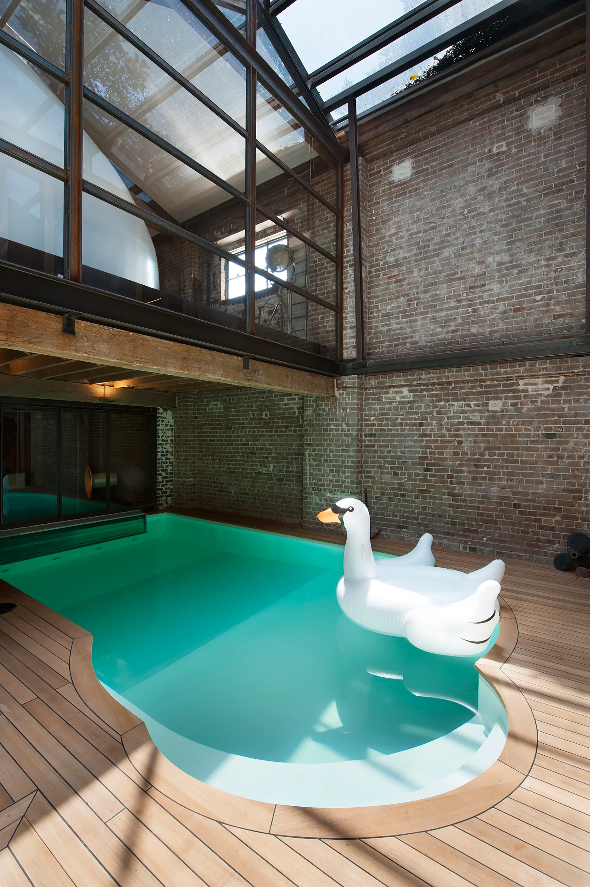An indoor swimming pool with turquoise water is surrounded by a wooden deck, exposed brick walls, and a large metal-framed glass skylight overhead.