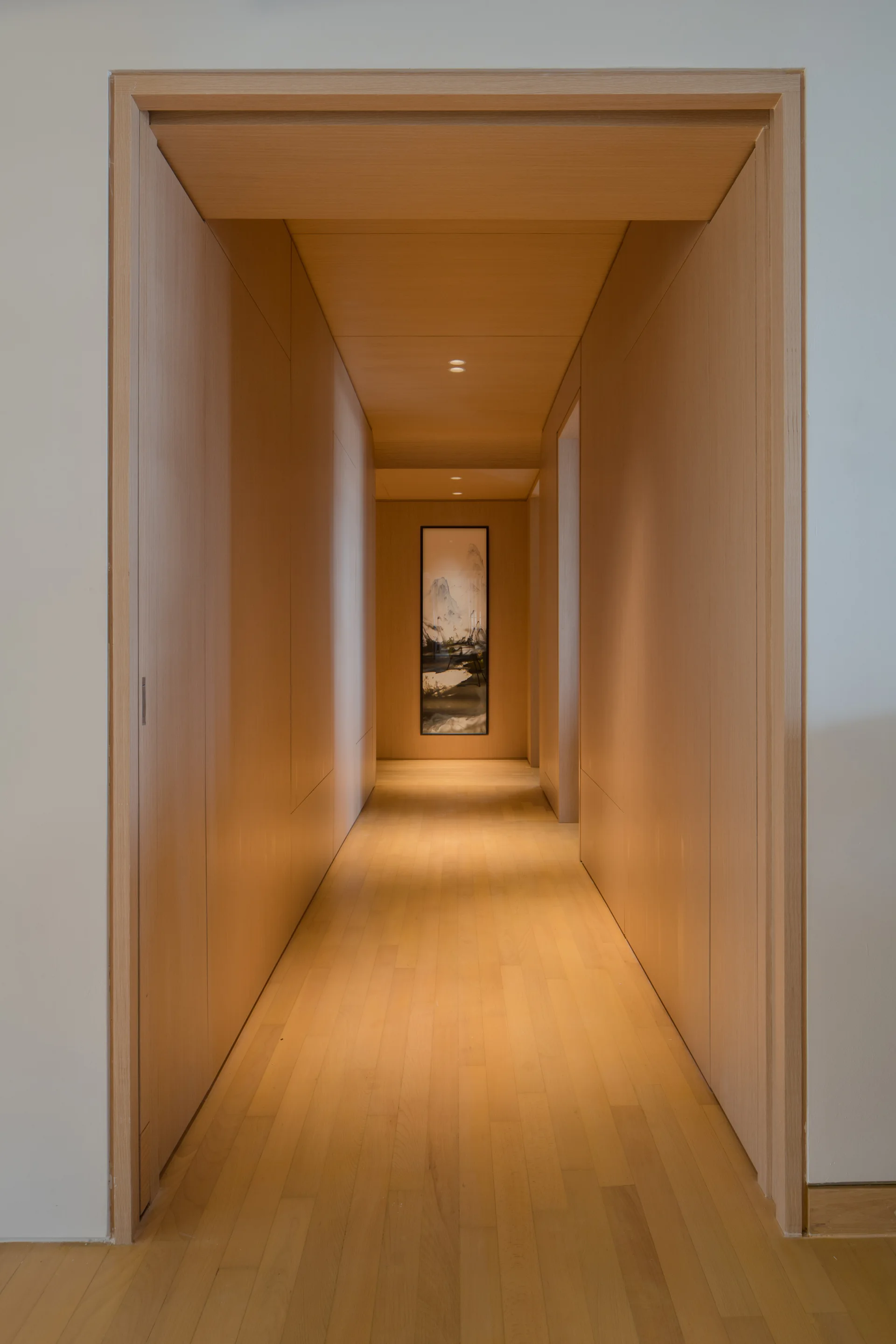 A symmetrical hallway lined with light wood panels features recessed ceiling spotlights and a framed vertical painting at the end.