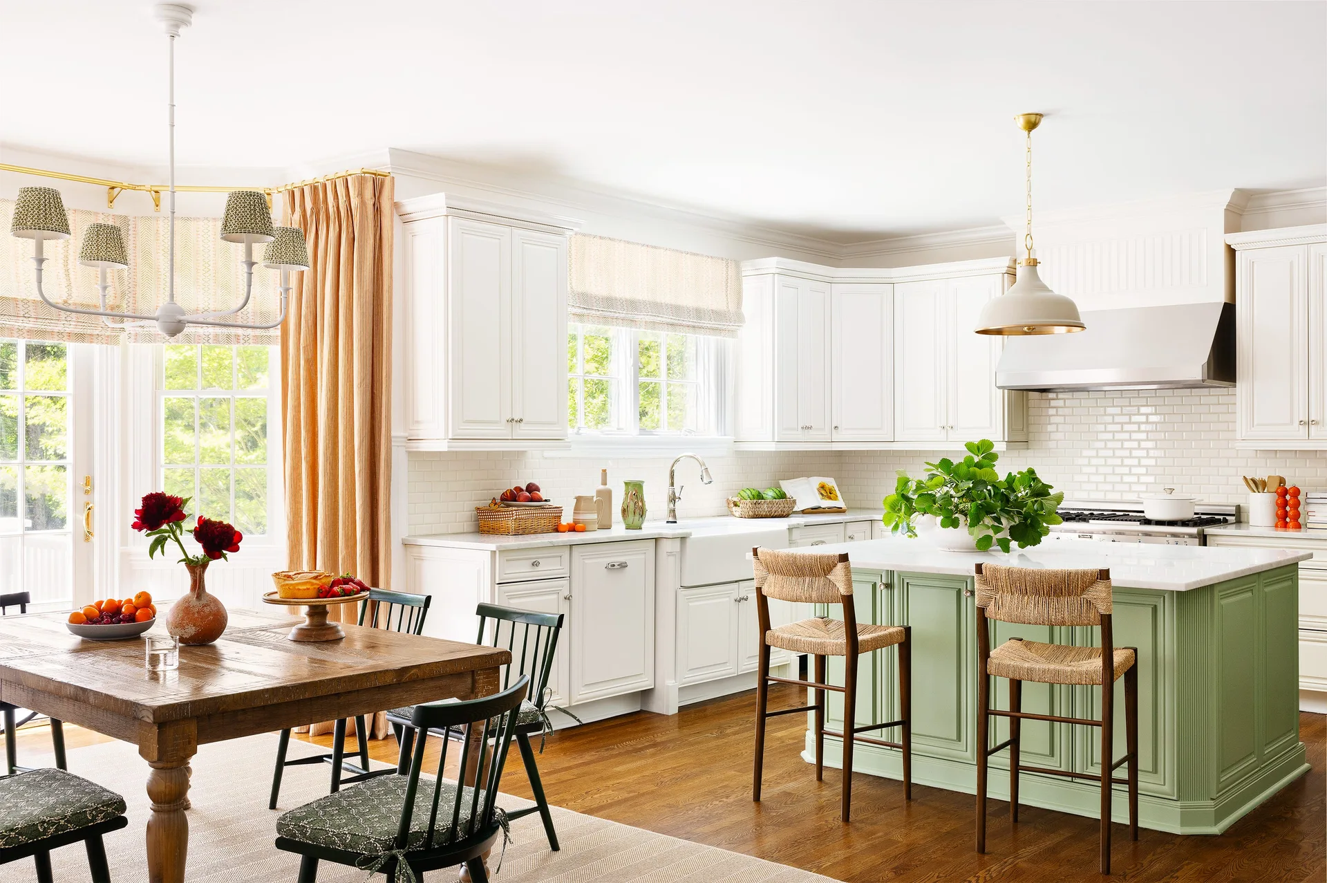 A bright kitchen with white cabinets, a sage green kitchen island with woven stools, and a wooden dining table.