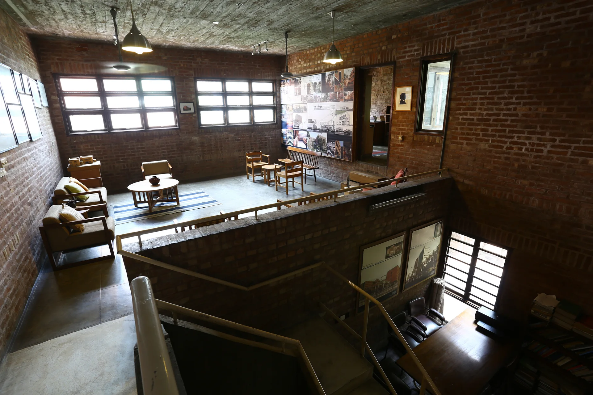An industrial split-level living space features exposed red brick walls, a raw concrete ceiling, wooden armchairs, and a staircase with a metal railing.
