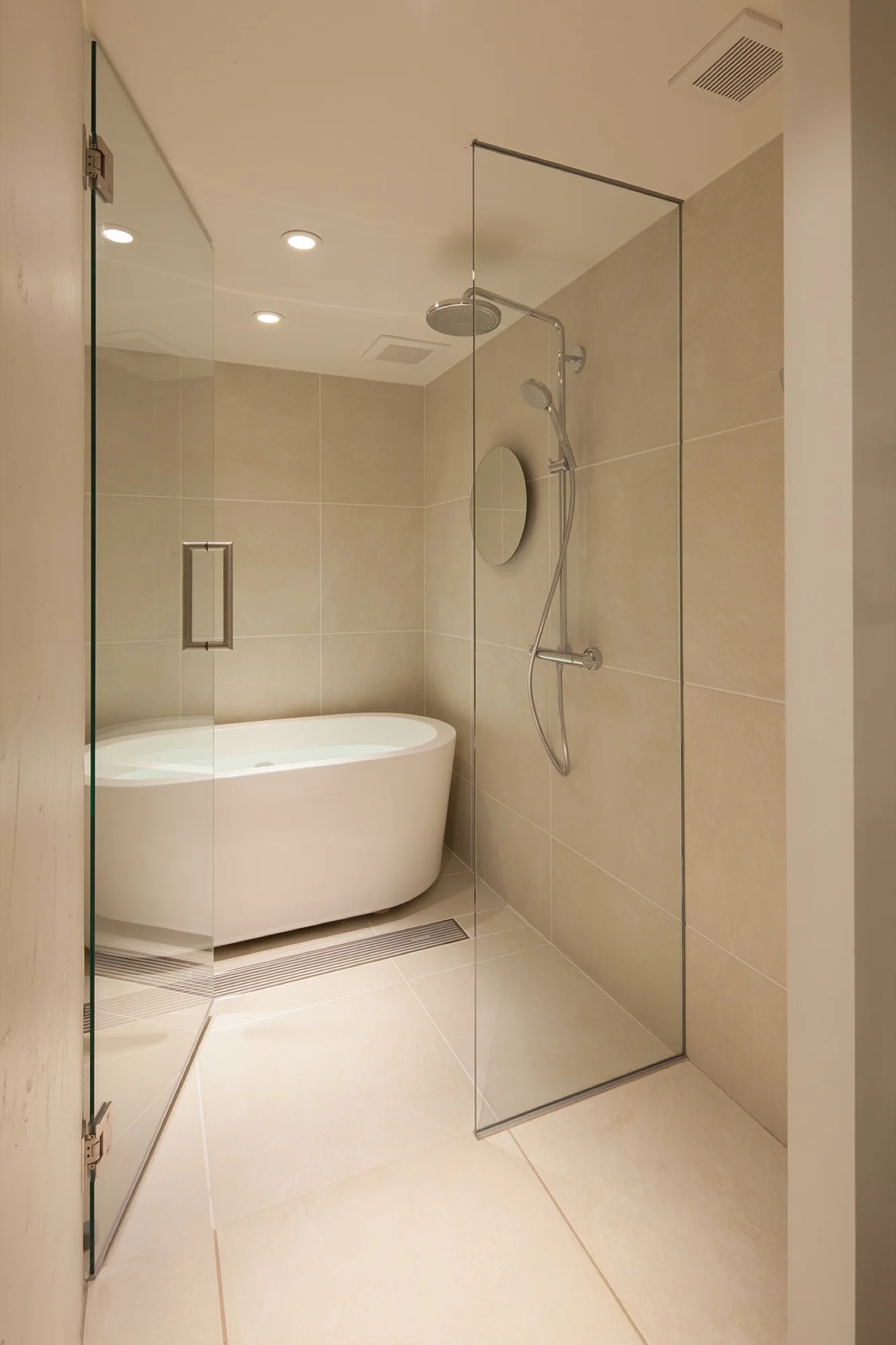 A modern beige-tiled bathroom featuring a white freestanding bathtub and a walk-in shower enclosed by clear glass panels.