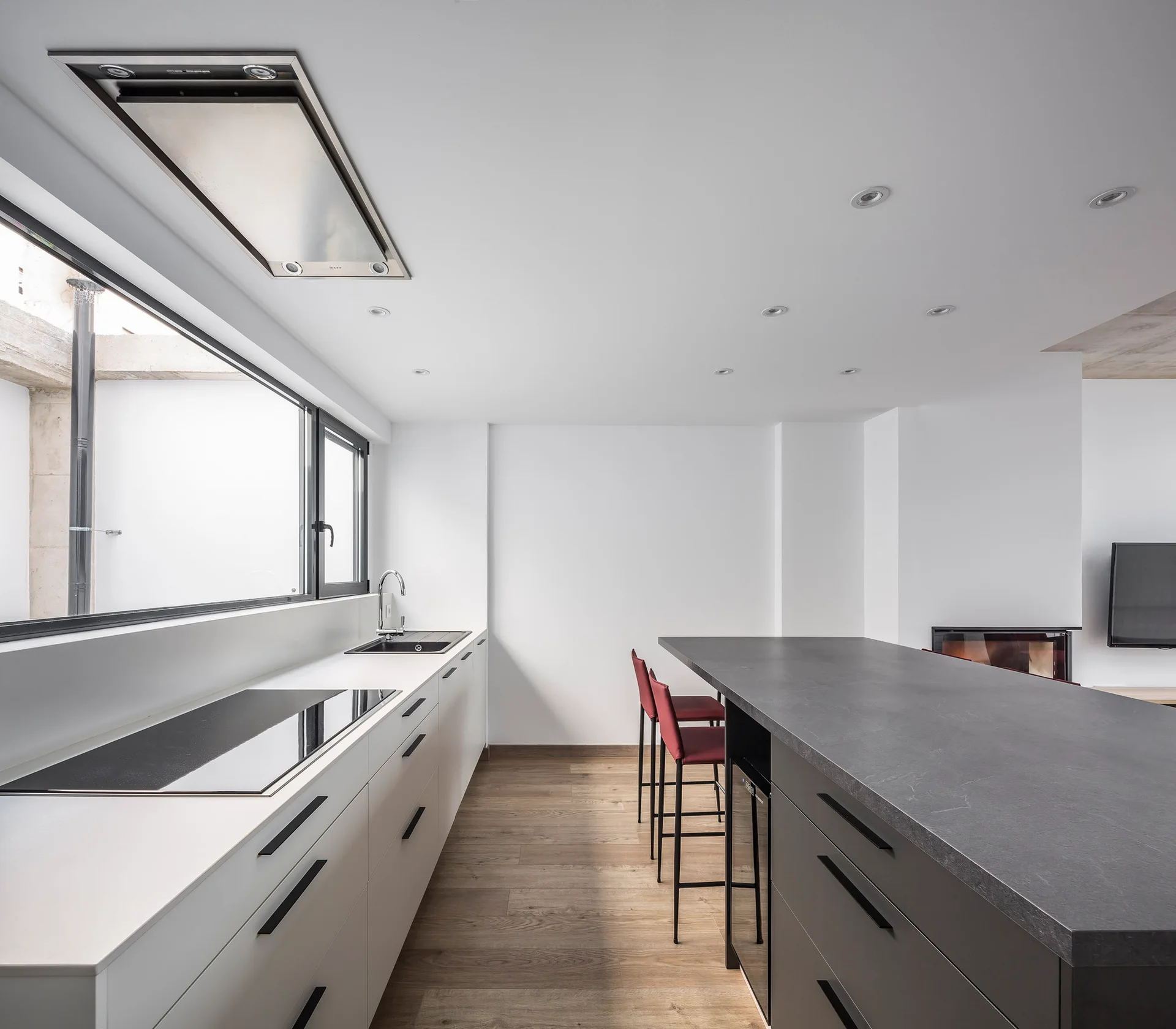 A minimalist kitchen features white cabinets with a black countertop cooktop, a large dark gray island with red bar stools, and warm wood flooring.