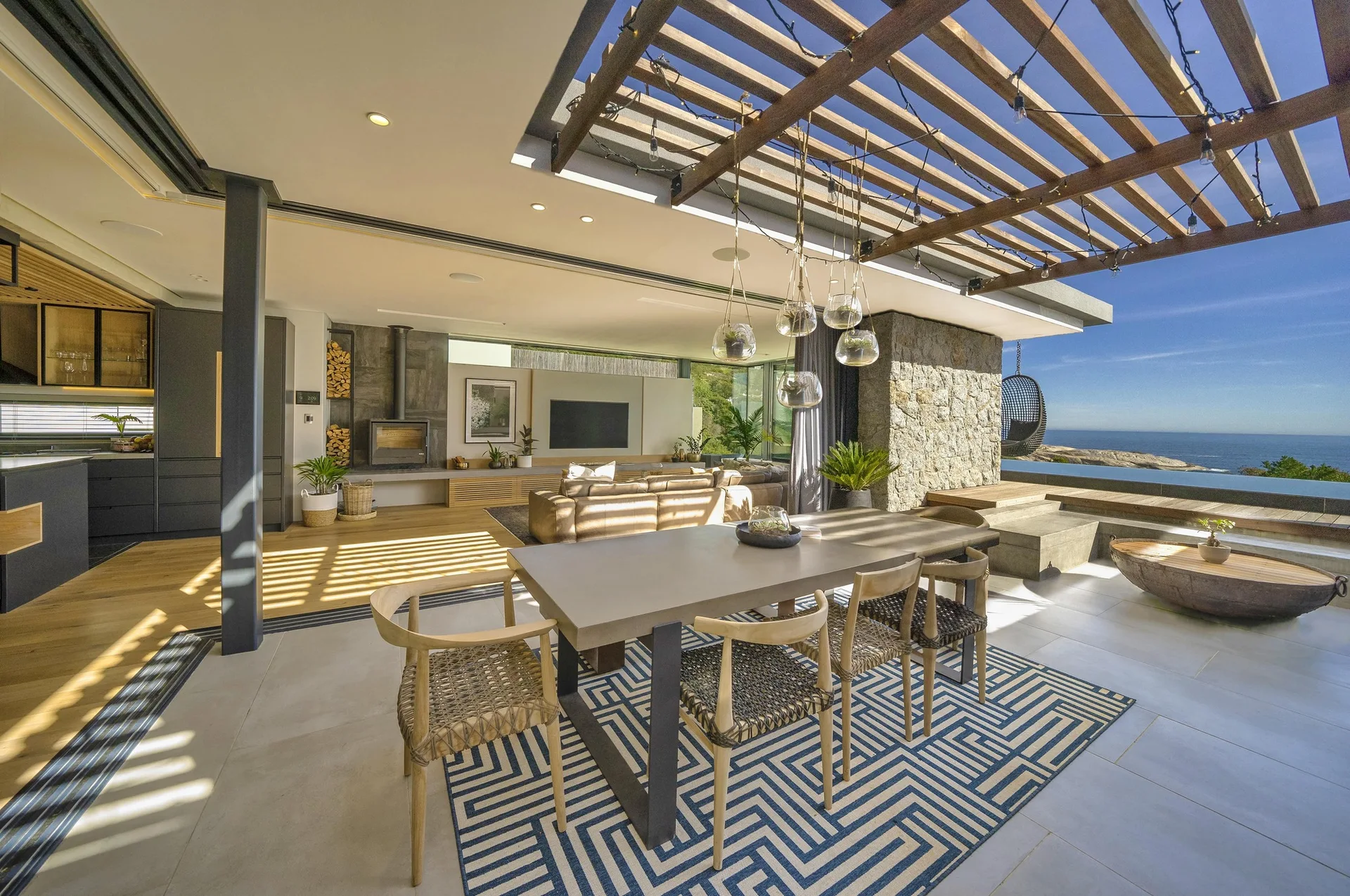 An outdoor patio dining table with woven chairs sits under a wooden pergola next to an open living area with an ocean view.
