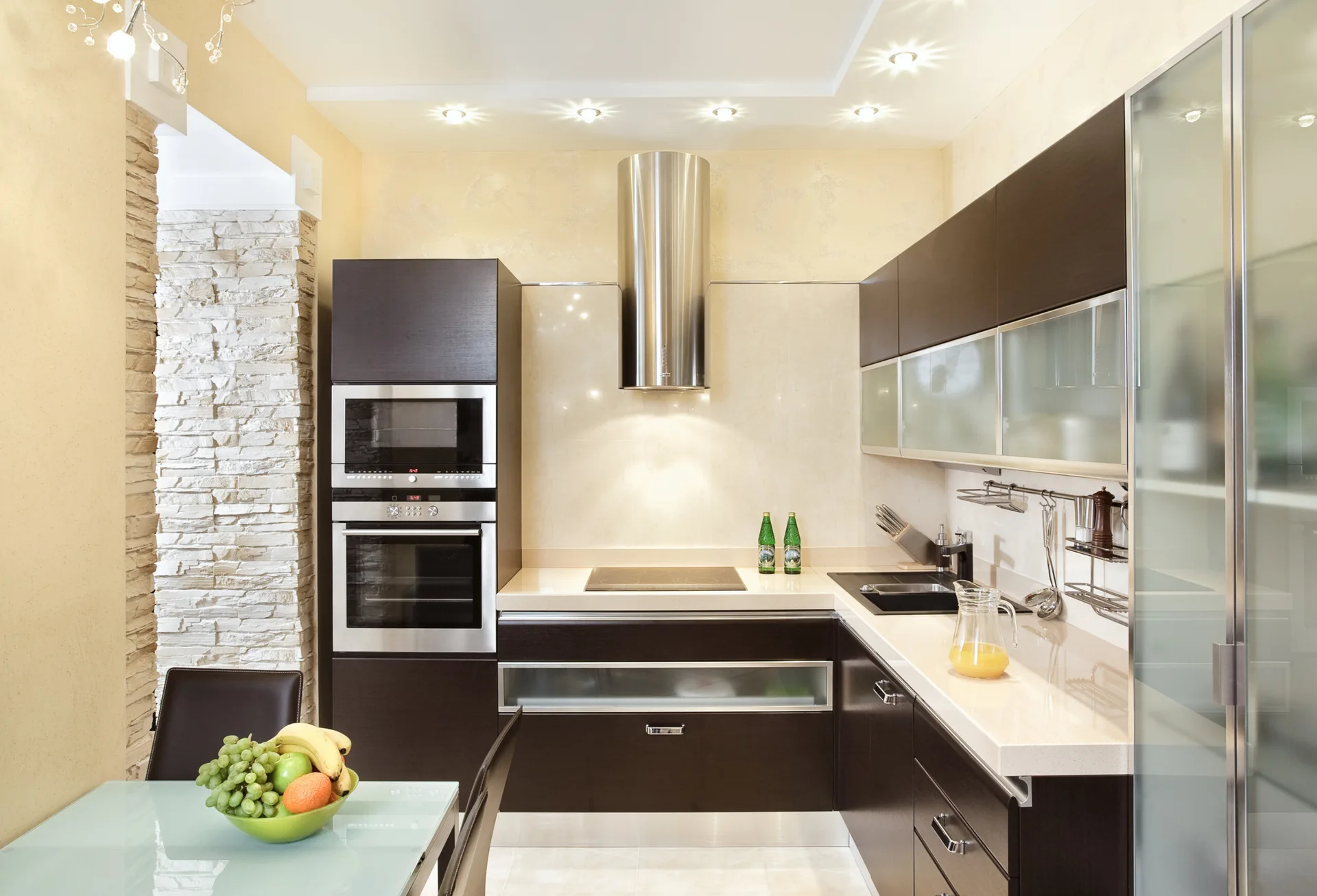 A modern kitchen features dark brown cabinetry, a stainless steel cylindrical range hood, cream countertops, and a textured stone accent wall.
