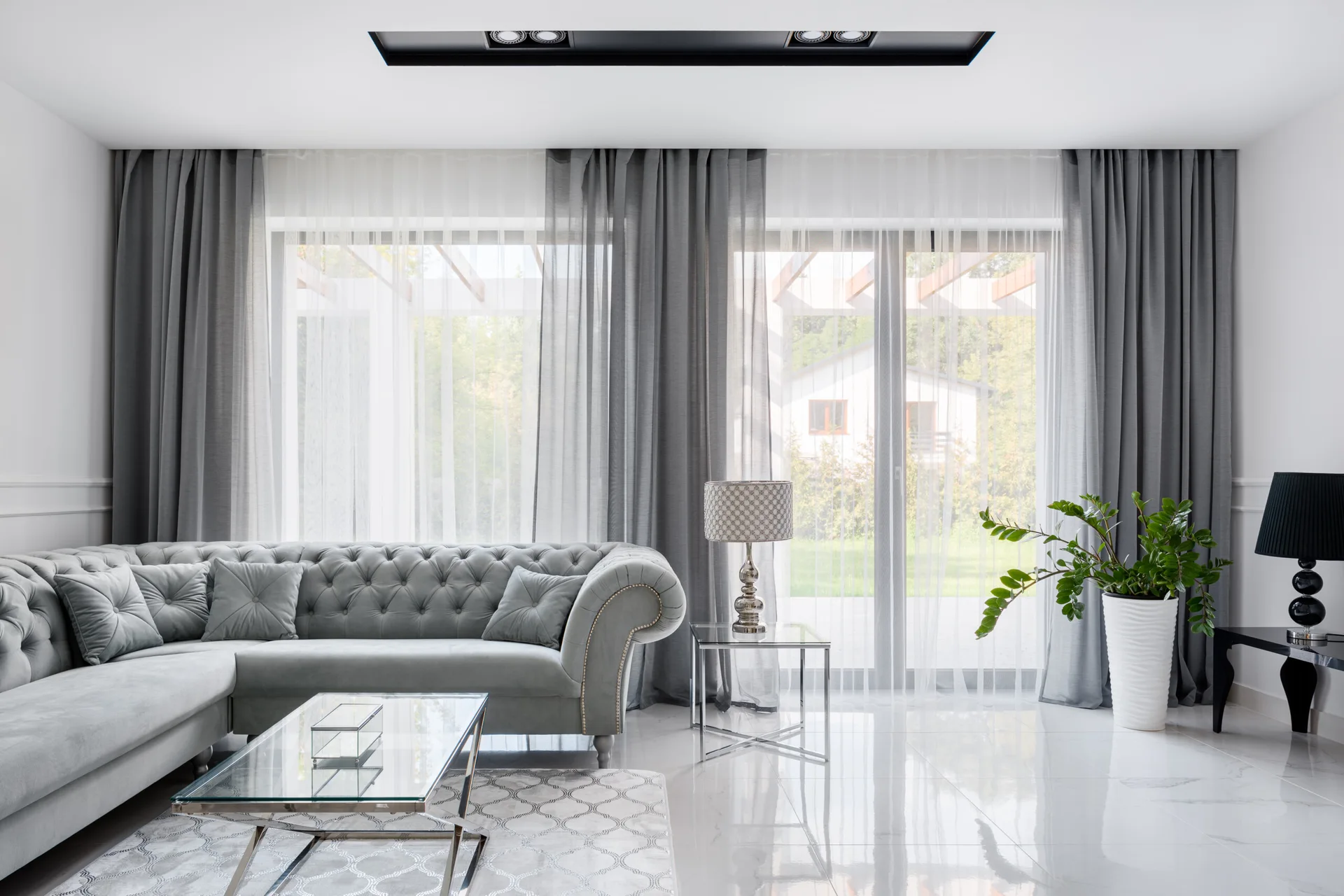 A modern living room features a grey tufted velvet sectional sofa, a glass coffee table, reflective white tile floors, and large windows with grey drapes.