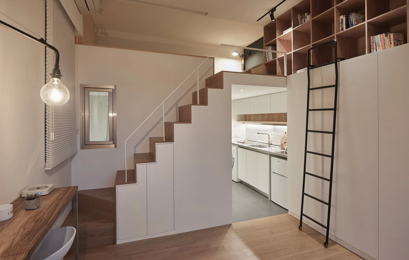 A minimalist studio apartment features a wooden staircase with built-in storage cabinets leading to a loft level above a small kitchen.