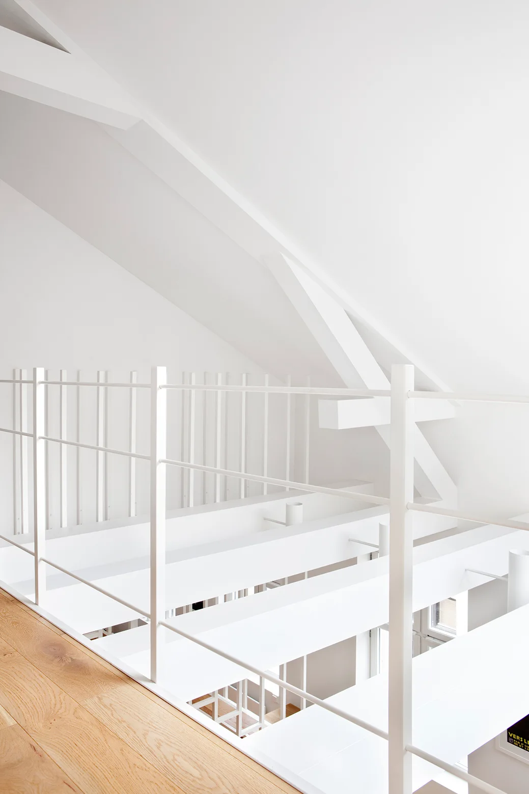 A modern mezzanine features a slender white metal railing alongside a light wood floor, beneath a sloped ceiling with painted structural beams.