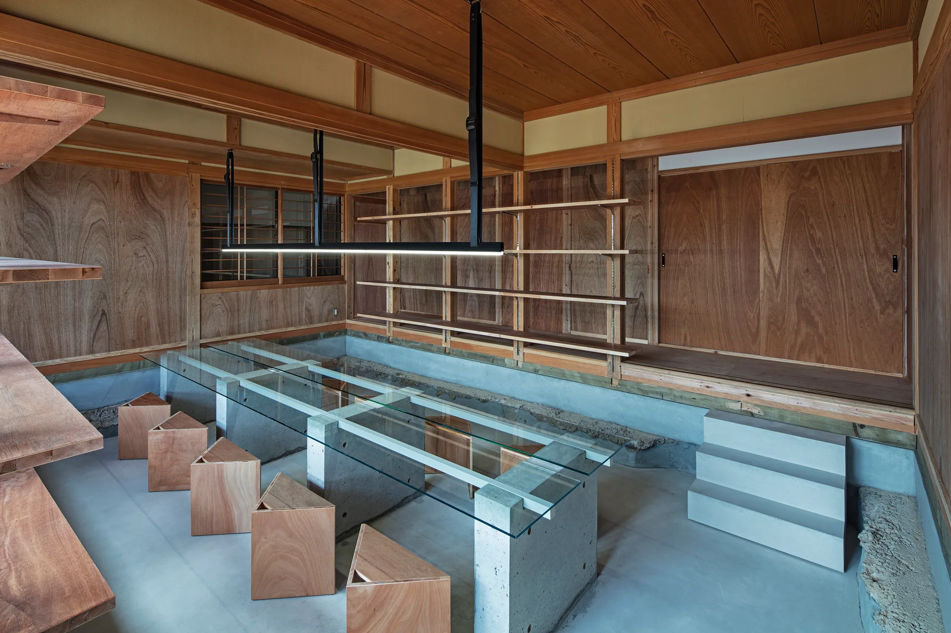 A minimalist workspace features a long glass-topped table with concrete block supports, triangular wooden stools, and wood-paneled walls.