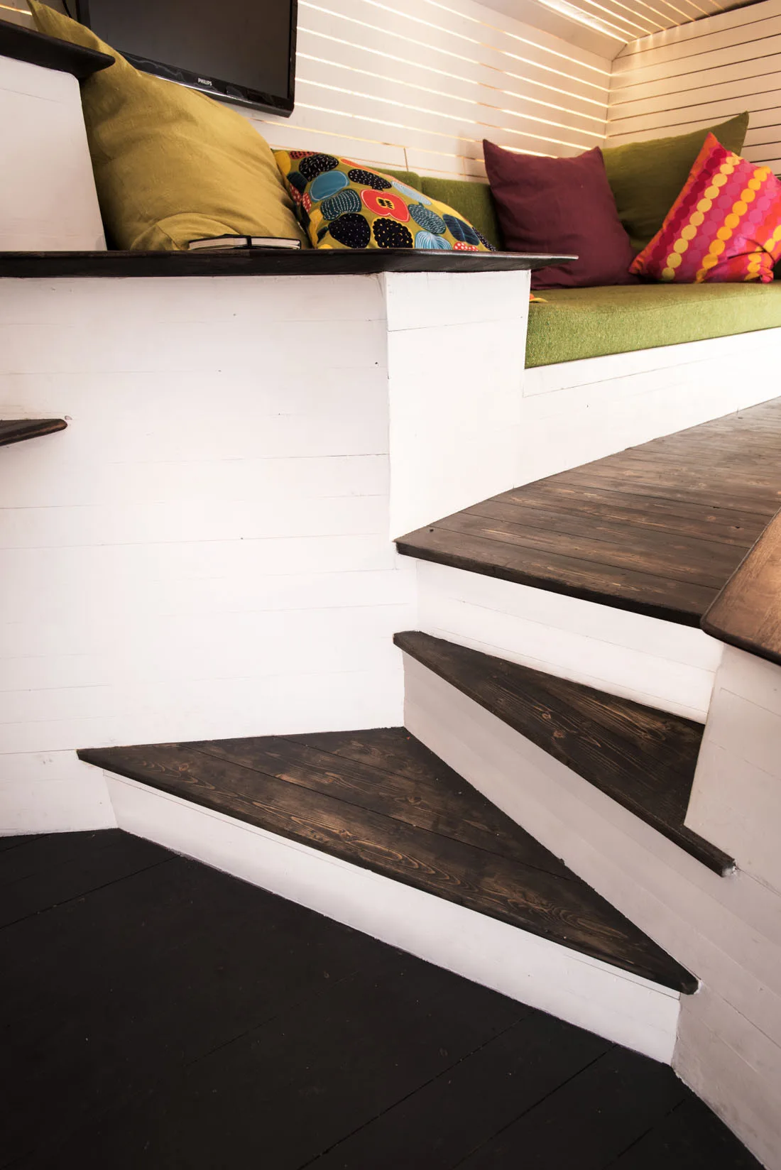 A built-in green cushioned bench sits next to a dark wood staircase with white painted wood-paneled walls and colorful throw pillows.
