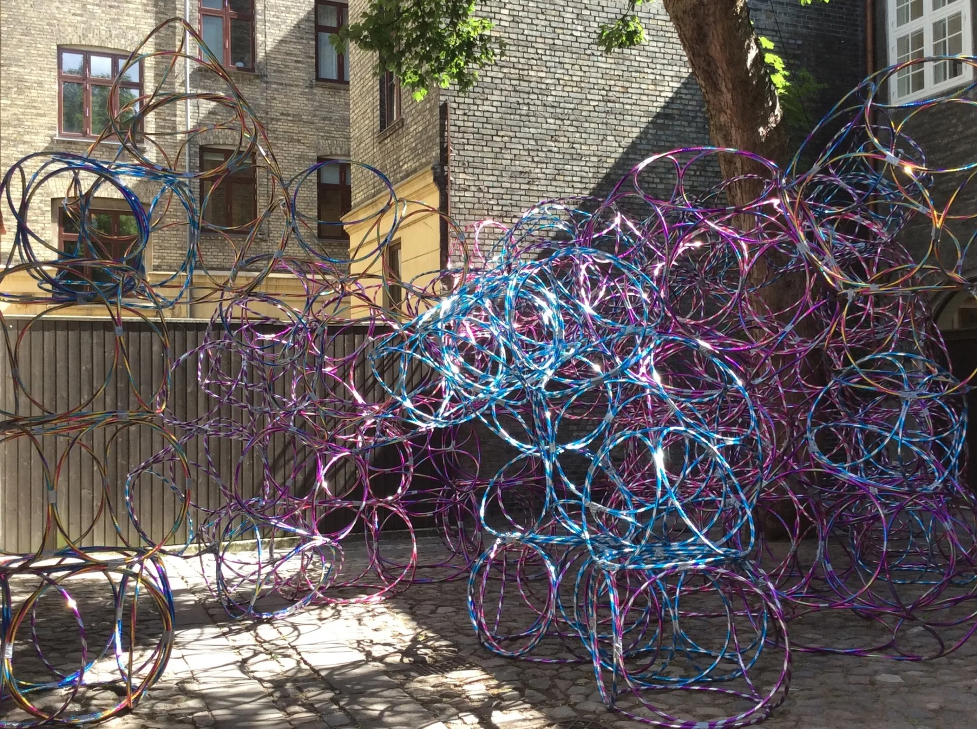 A large abstract sculpture made of overlapping iridescent blue and purple hoops stands in a cobblestone courtyard with brick building facades.