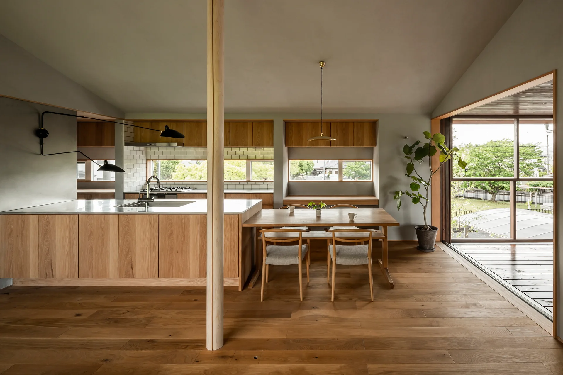 A minimalist kitchen and dining area with warm wood cabinets, a wooden support pillar, light gray walls, and sliding glass patio doors.