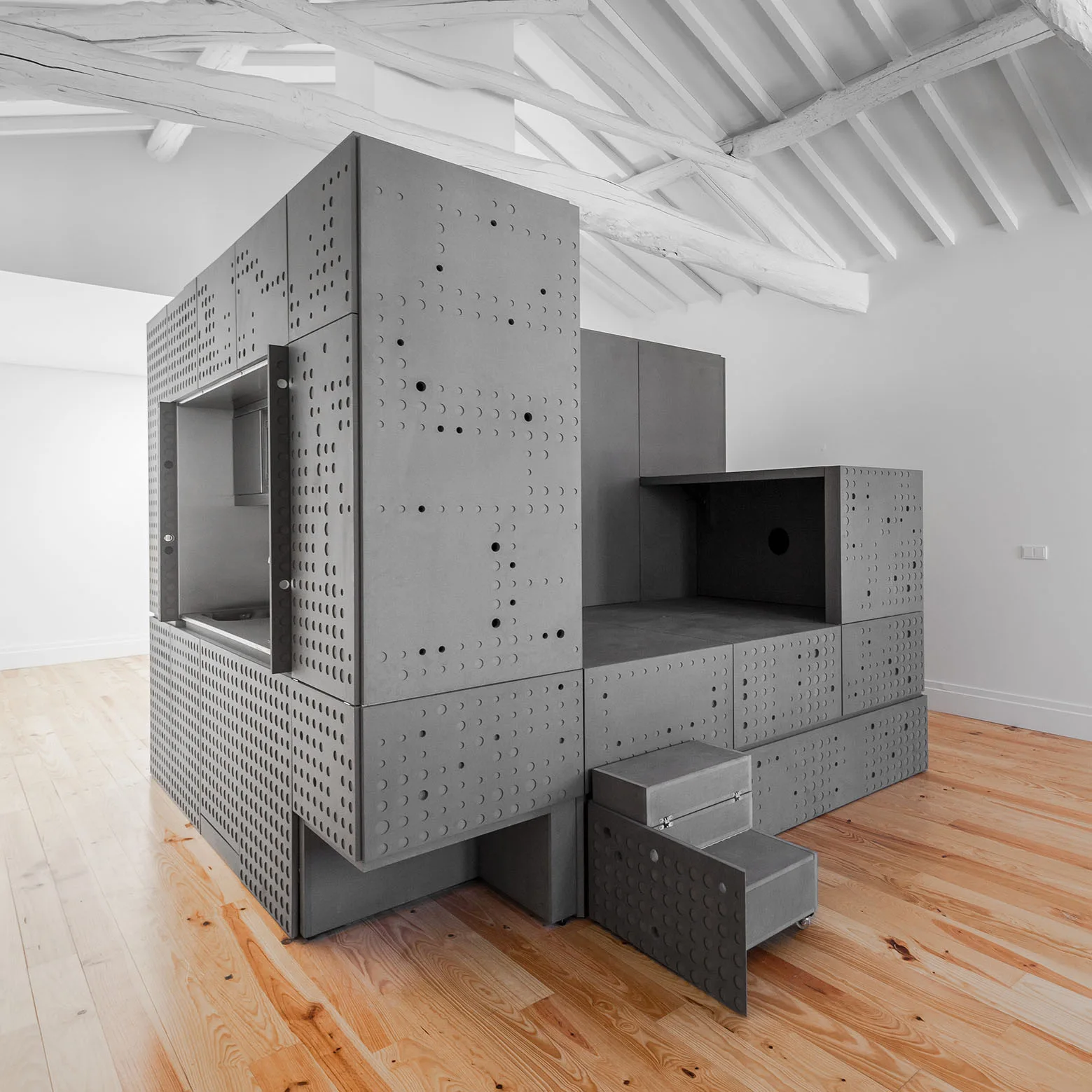 A large grey modular storage unit with perforated panels stands in an open room with light wood floors and white exposed ceiling beams.