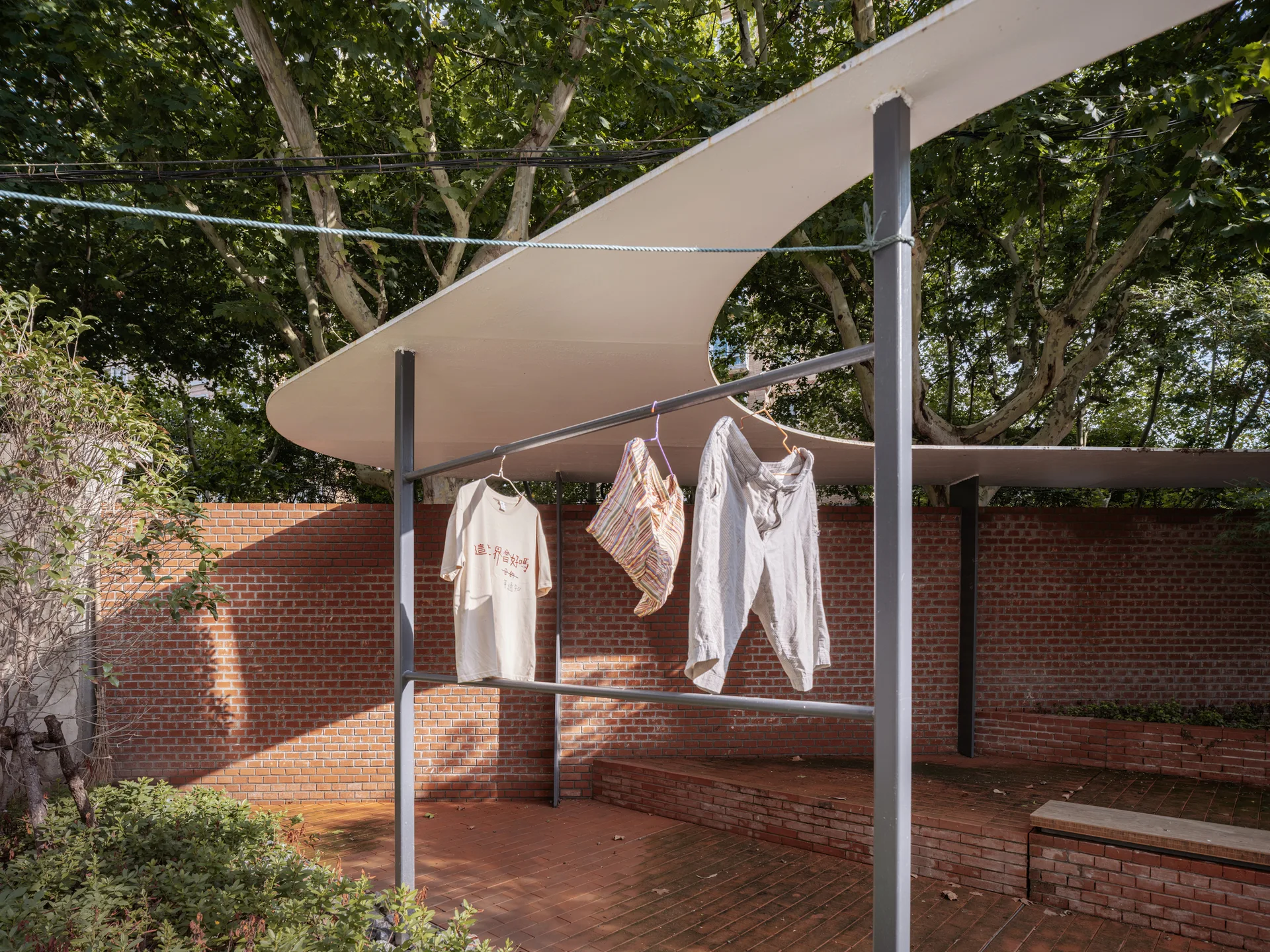 A modern outdoor courtyard featuring a curved white canopy structure, a red brick wall, and a grey metal drying rack on brick pavers.