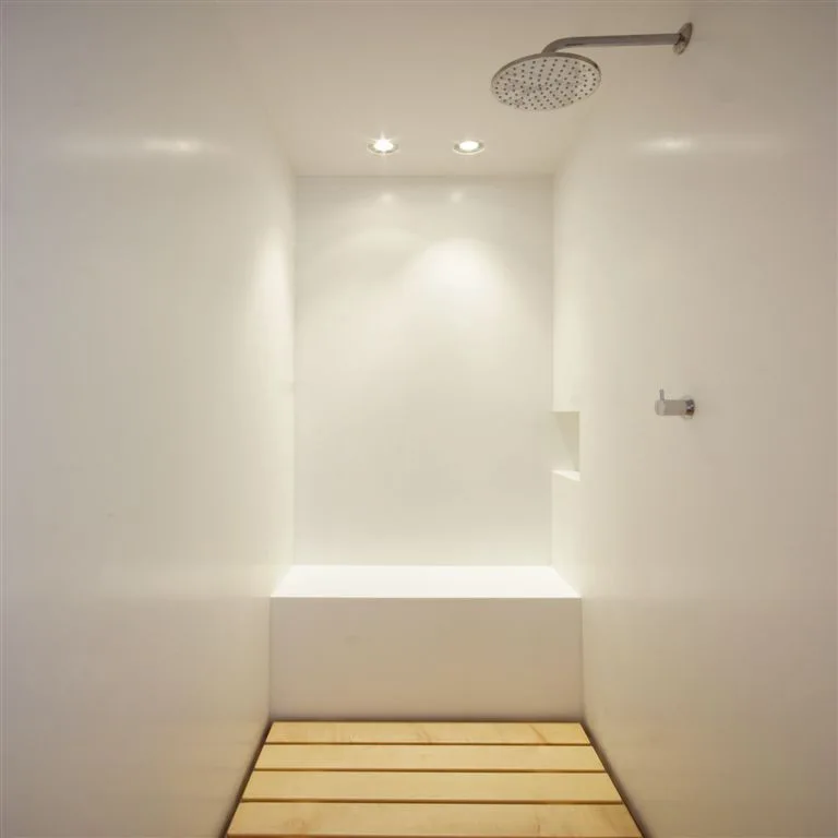 A minimalist white shower enclosure features a built-in bench, an overhead rain shower head, and a light wood slatted floor insert.