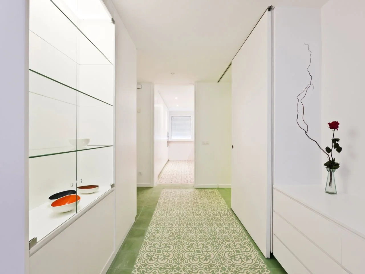 A minimalist white hallway featuring a green patterned tiled floor, built-in glass display shelving, and sleek handleless white cabinetry.