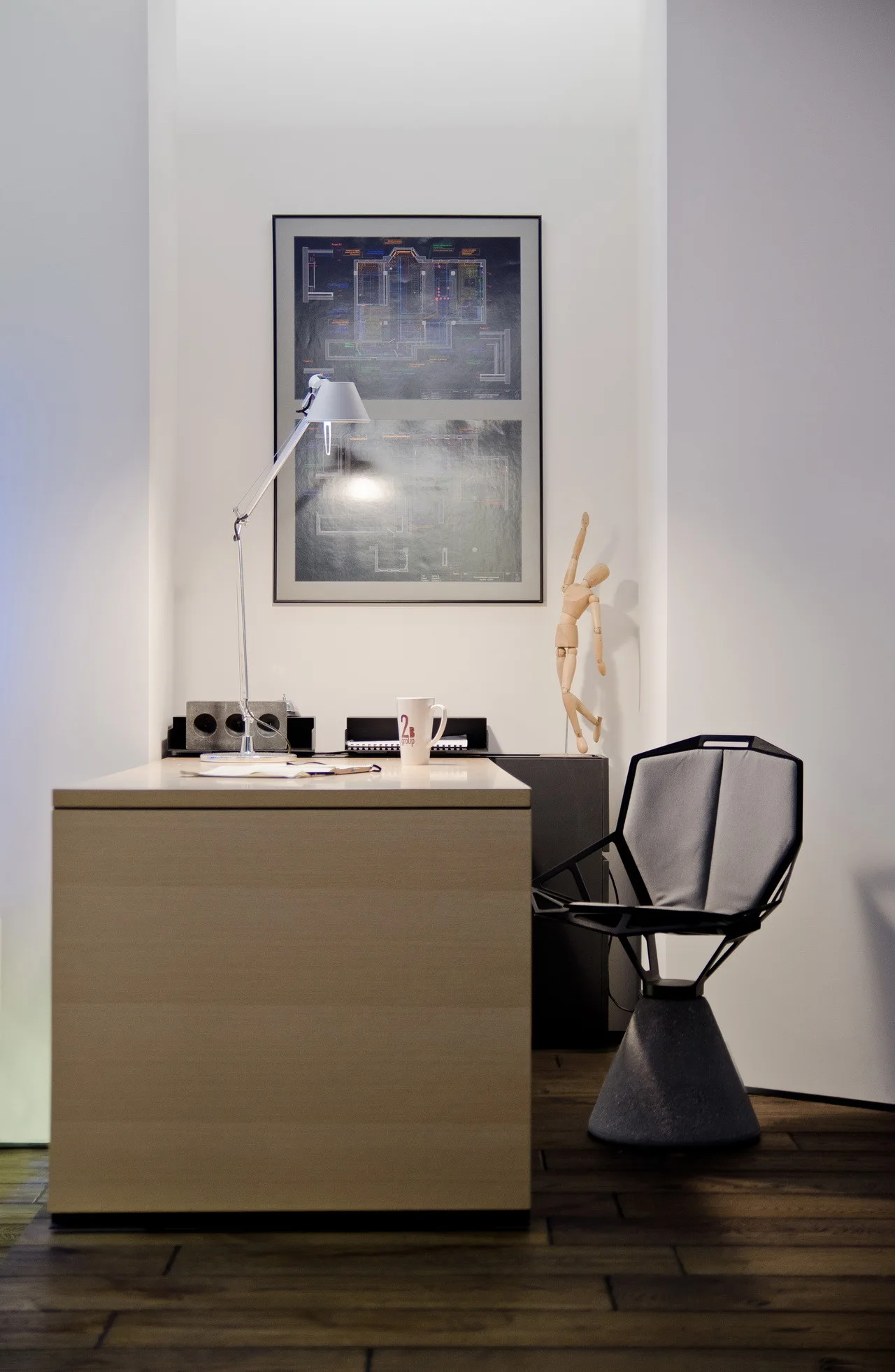 A light wood desk and sculptural gray chair sit in a white wall alcove beneath a framed architectural blueprint print.
