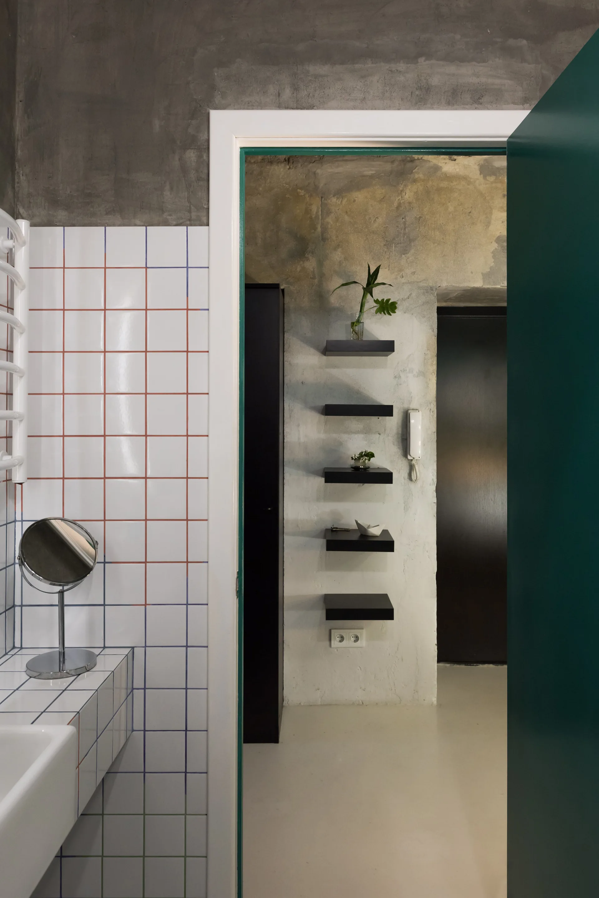 A bathroom with grid-patterned white tiles and a table mirror looks into a hallway with raw concrete walls and small black floating shelves.