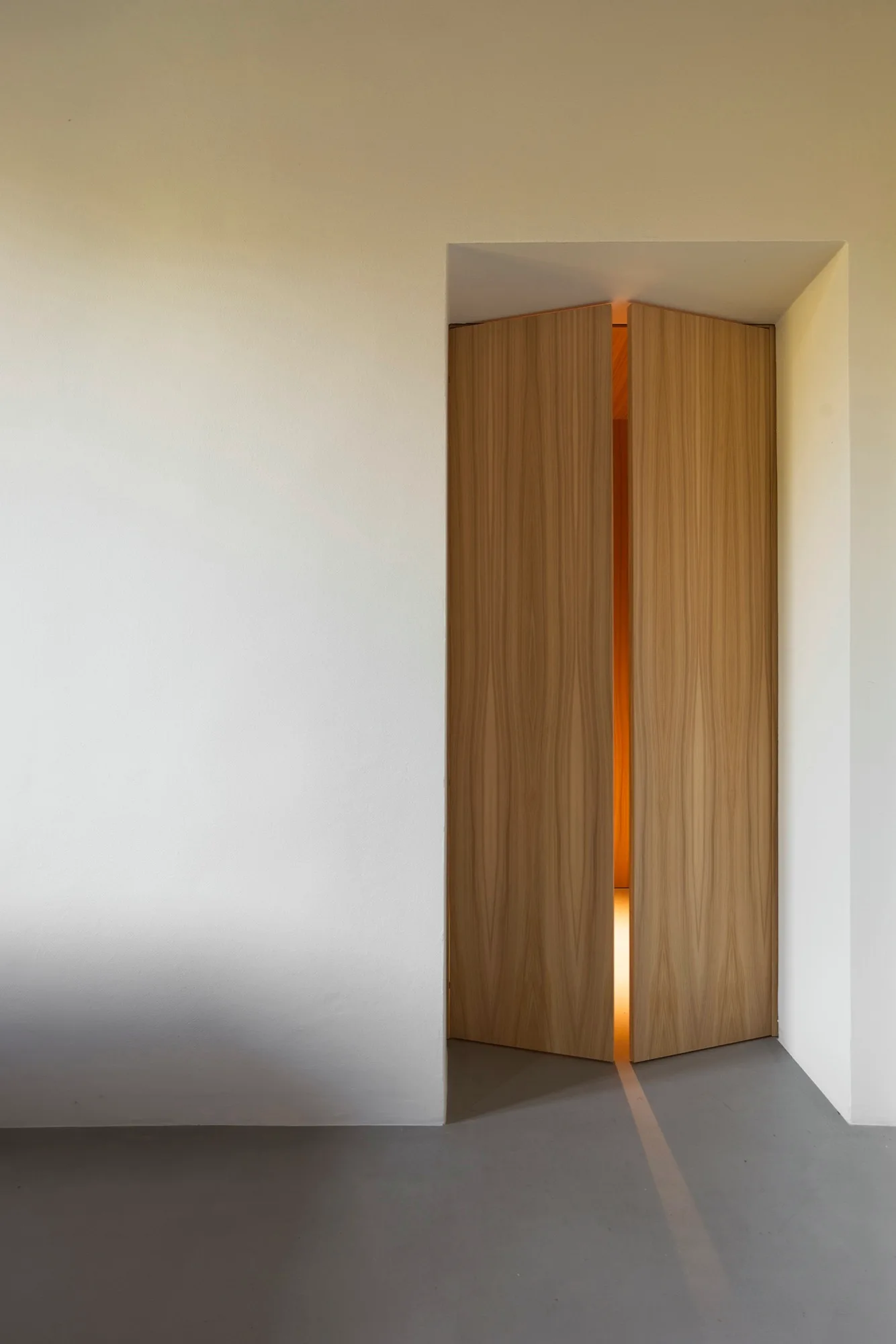 Slightly ajar light wood double doors set into a minimalist white recessed wall with warm light glowing through the opening.