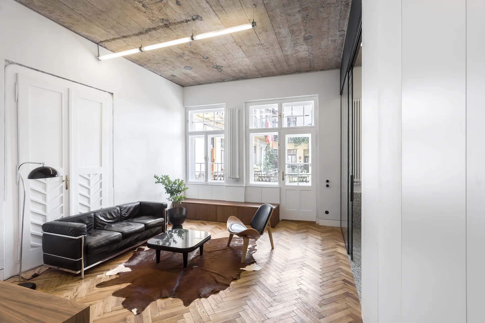 A modern living room featuring an exposed concrete ceiling, black leather sofa, and cowhide rug over herringbone wood flooring.