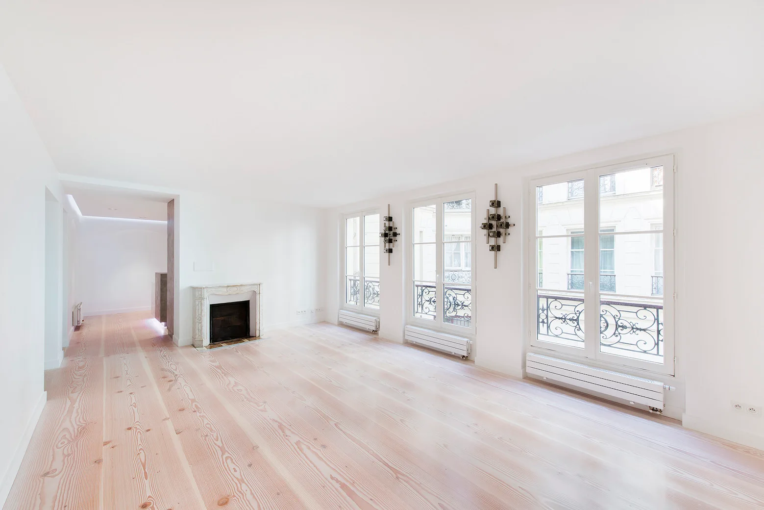 An empty white room features wide-plank light wood flooring, a stone mantel fireplace, and three large French windows with black wrought-iron railings.