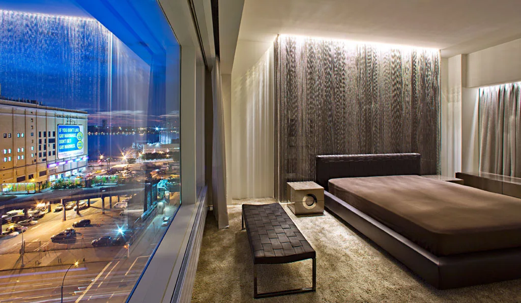 A modern bedroom with a low-profile bed, a woven leather bench, and a metallic chain curtain wall illuminated by glowing cove lighting.