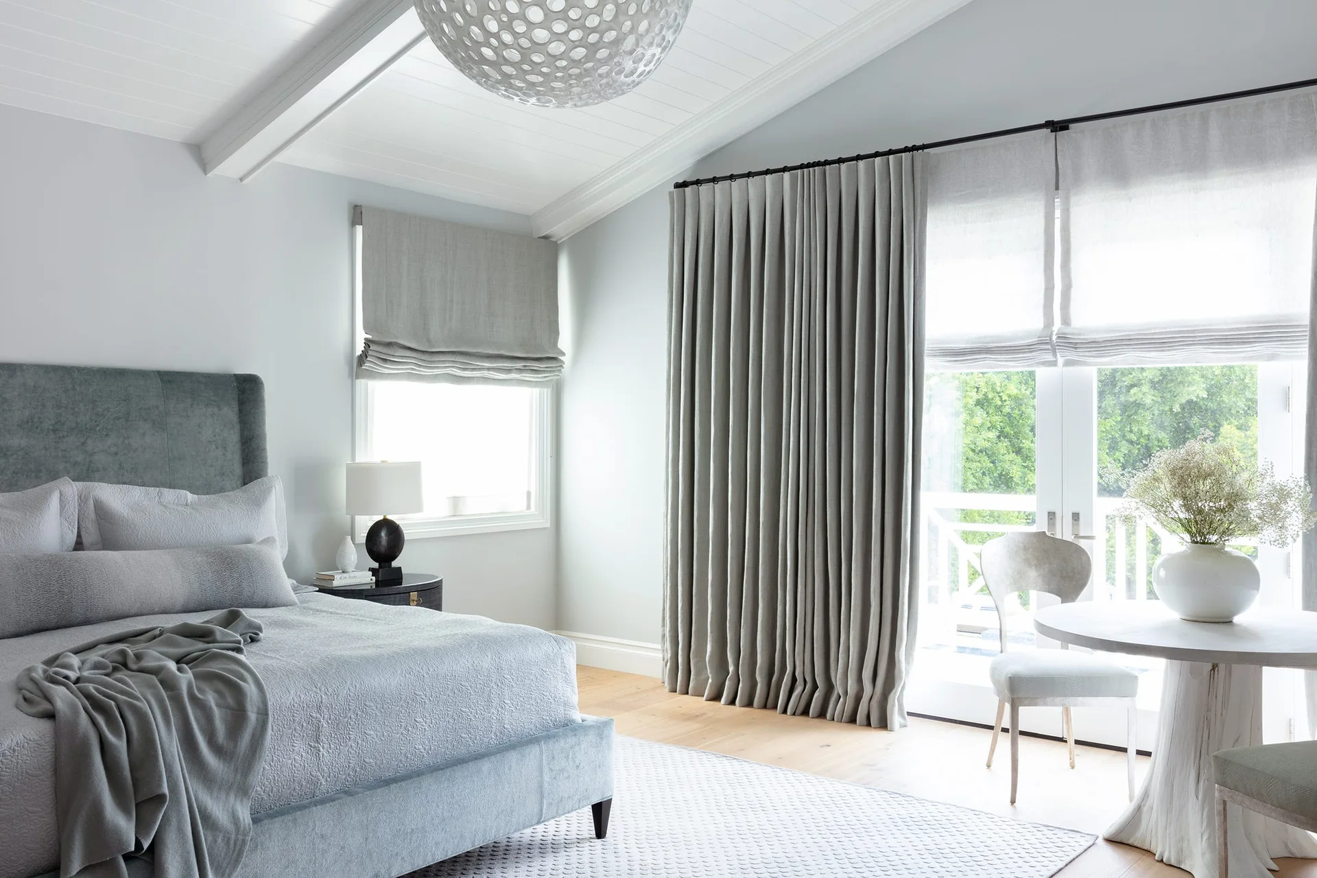A light grey bedroom features an upholstered bed, a vaulted ceiling with white beams, a round pedestal table, and long grey curtains by glass doors.